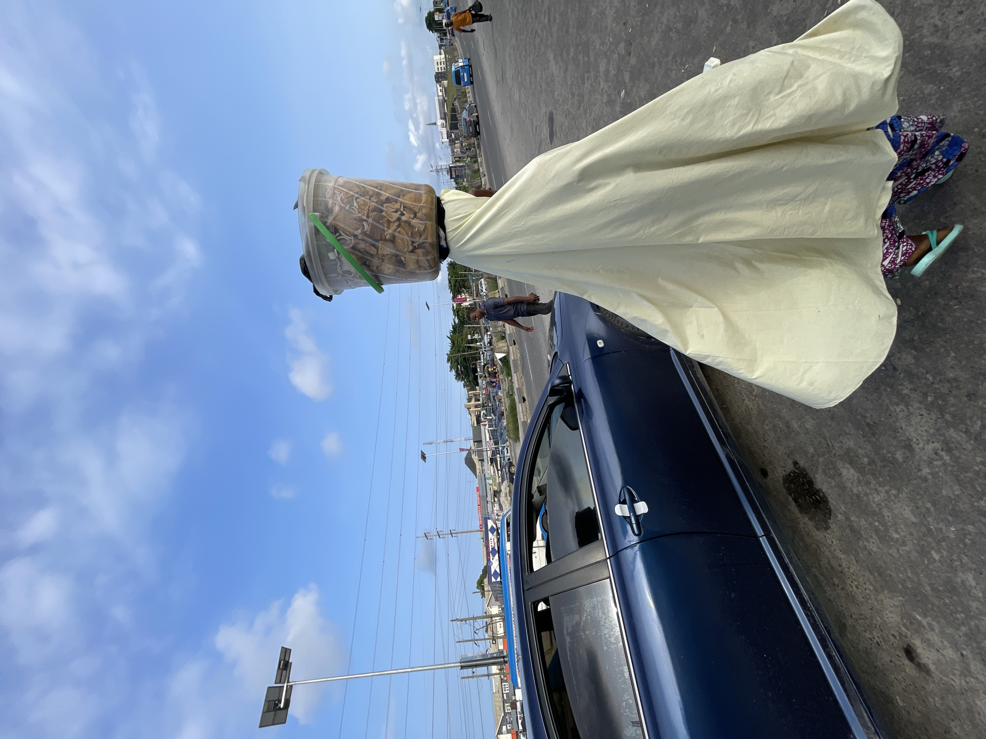 In Lekki, Nigeria, a woman is captured walking with a large plastic bucket filled with food items balanced on her head, draped in a pale yellow covering. To her left, a dark blue sedan is parked. The scene unfolds on a paved road, with a man walking in the background and other vehicles visible further down the street. Power lines and streetlights dot the sky, which is partly cloudy. The overall environment suggests an outdoor urban setting.