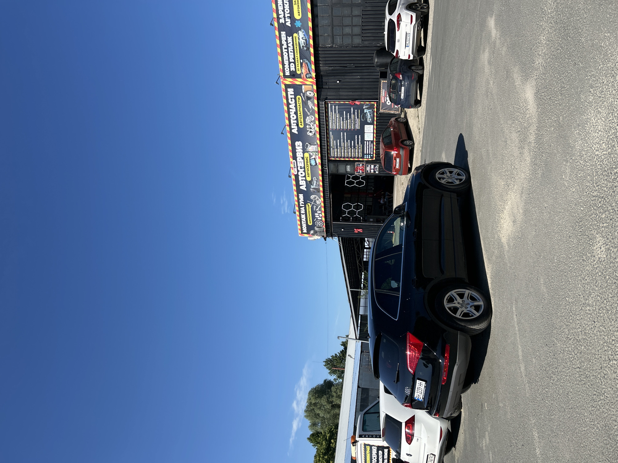 An outdoor scene shows a row of cars parked in front of a building with a large banner. The banner, in Ukrainian, advertises "Auto Parts" and "Auto Service," with smaller text listing services like tire mounting, computer diagnostics, and air conditioning repair. The building appears to be a garage or service station, with open bays visible. The cars are a black Audi SUV, a white Volkswagen hatchback, a red compact car, a dark blue hatchback, and a white Hyundai SUV. The sky is clear blue. The ground is asphalt. The location is the city of Nesebar, Bulgaria.