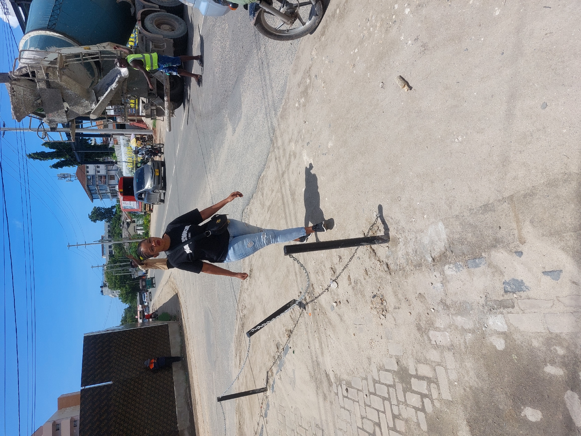 This image depicts an urban street scene in Dar es-Salaam, Tanzania, on a bright, sunny day, indicated by the clear blue sky and strong shadows.
The main subject is a woman standing on a paved sidewalk, posing for the camera. She is wearing a black t-shirt with the word "COMPLEX" and possibly "EST. 1997" visible on it, light blue distressed jeans, and dark sneakers. She carries a black shoulder bag. She is positioned next to a chain barrier with black posts, creating a division between the sidewalk and a dirtier section of ground.
In the background, on the street to the left, a large cement mixer truck is visible. A man wearing a bright yellow high-visibility vest and shorts is standing next to the truck. Chinese characters (e.g., "黄色" which means yellow, and a partial "1989" or "1988") and some English text ("MADE") are visible on the truck's side. Further down the road, parked motorcycles and a black car (with a license plate starting "T...D") are seen. Beyond these, there are various buildings, some with multiple stories, and green trees. Power lines crisscross the upper left portion of the image.
On the far left, a structure covered with a brown/black material, repeatedly marked "ZEBRA FORM," appears to be a construction hoarding or wall. Another person in work attire is visible near this structure. The ground consists of a mix of concrete, asphalt, dirt, and sections of brick pavers. The overall scene suggests a busy area with ongoing construction or development.