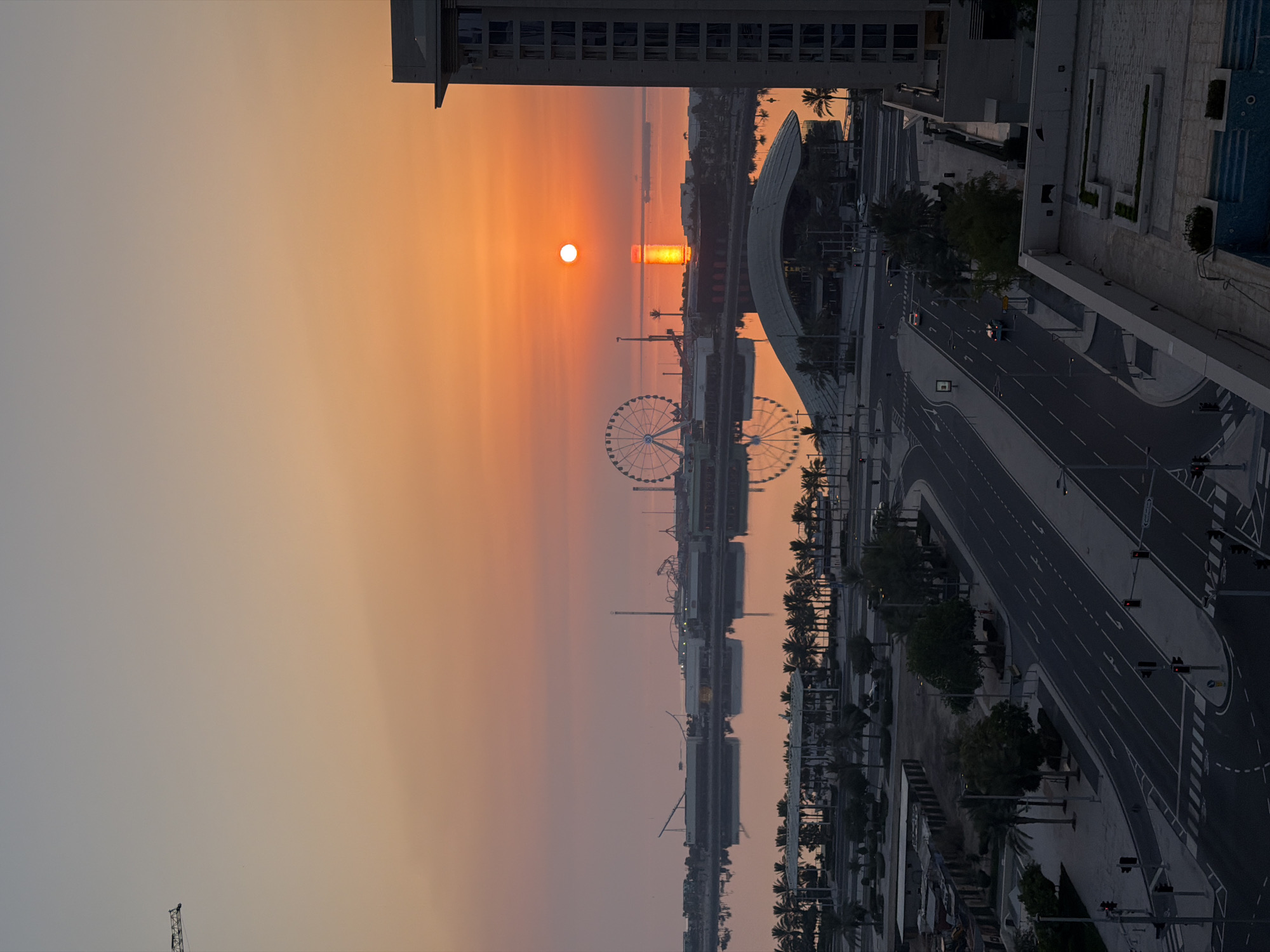 The image depicts a sunrise over a city skyline, likely in a coastal area, given the presence of a large body of water. The sun, a bright orange orb, is positioned low on the horizon, casting a warm glow across the sky which transitions from a pale yellow to orange and then to a soft pink hue. A prominent Ferris wheel is visible in the mid-ground, silhouetted against the vibrant sky, along with other amusement park structures and buildings. The water reflects the colors of the sunset, creating a mirrored effect. In the foreground, a multi-lane road curves through the scene, with traffic lights and pedestrian crossings. Palm trees line the roadside and are also scattered throughout the urban landscape. To the right, a tall, modern building stands, its windows catching the light. There are no people clearly visible in the image, but there are several vehicles on the road and a few structures that suggest human activity and development. The overall scene conveys a sense of a waking city at the beginning of a new day.