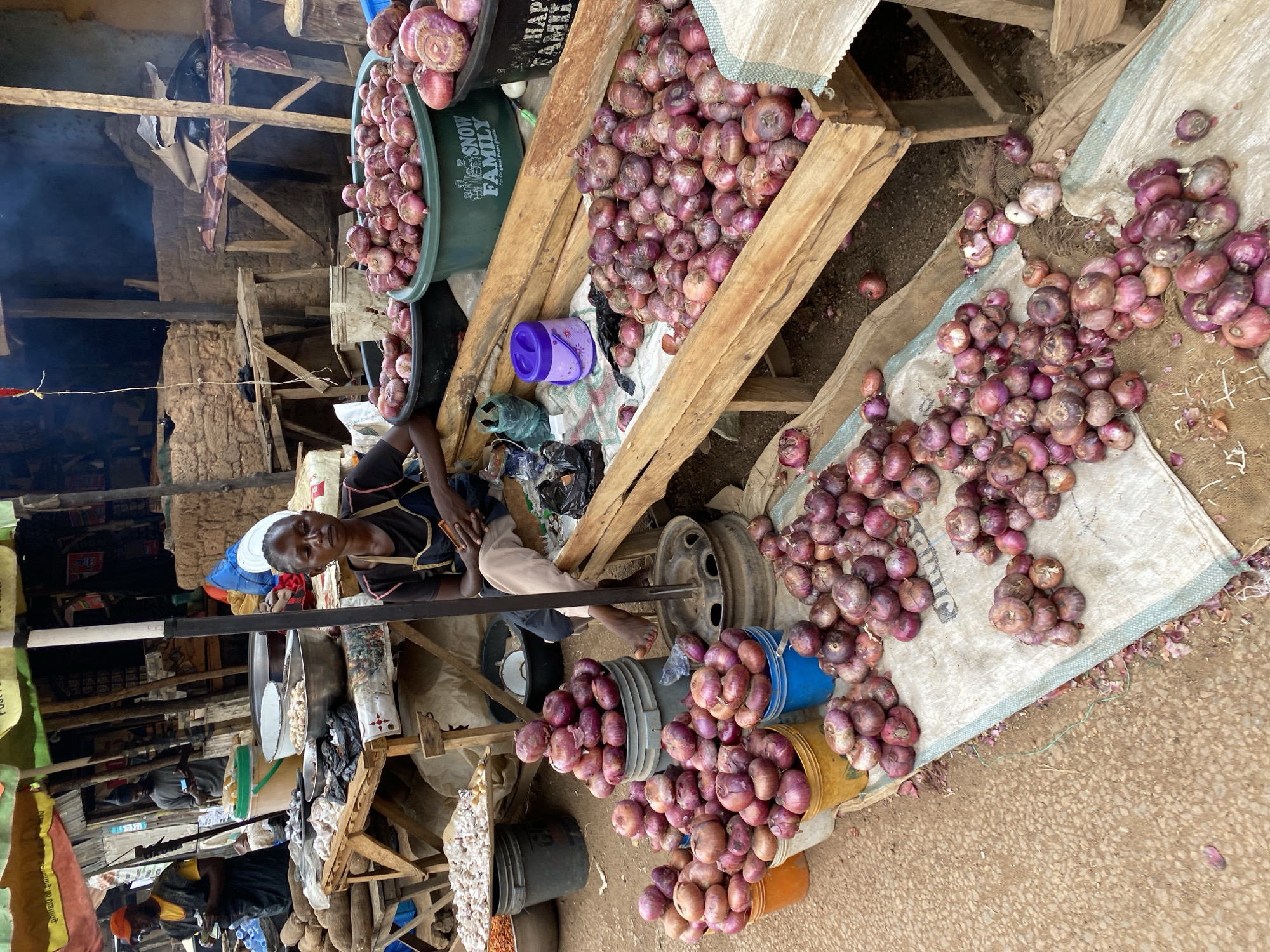 In the city of Bukuru, Nigeria, a woman sits at an outdoor market stall laden with produce. She is wearing a dark t-shirt and light-colored pants, and is seated on a wooden structure. The stall is filled with numerous piles and buckets of red onions. A large wooden crate overflowing with onions is positioned in front of her. Other market stalls and vendors are visible in the background, with some people milling about. The market appears to be open-air with makeshift structures and stalls. The overall scene depicts a busy market day in Bukuru.