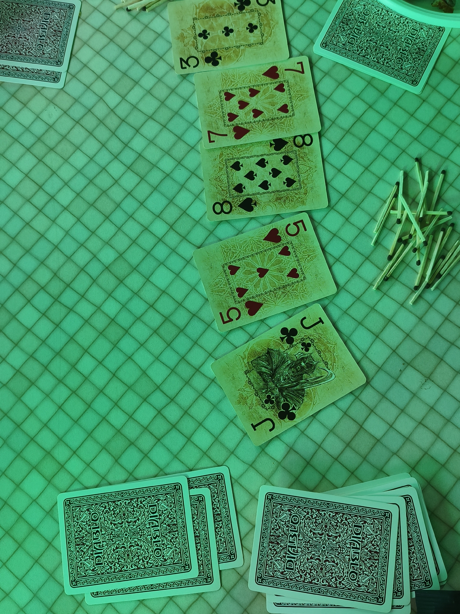 The image displays a collection of playing cards and a pile of matches arranged on a green checkered surface. Several cards are face up, showing their numerical and suit markings, including a 3 of clubs, a 7 of hearts, an 8 of spades, a 5 of hearts, and a Jack of clubs. Other cards are face down, their backs featuring an intricate "DIABLO" design. The matches are clustered together in a pile. The lighting casts a green hue over the entire scene. This scene is not identifiable as being in Chaikovsky, Russia.