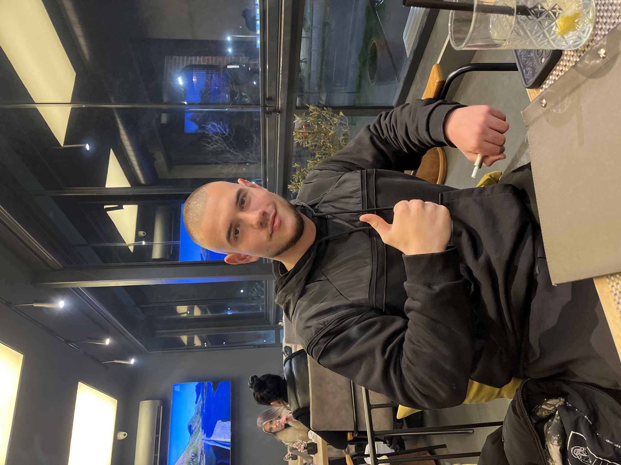 The image shows a man sitting at a table in a cafe or restaurant. He is fair-skinned and has a shaved head. He is wearing a black hooded jacket. He is holding a thin, white cigarette between his fingers on his right hand and is giving a thumbs up with his left hand. On the table is a glass with a beverage and a straw. A cellular phone also rests on the table. The setting is a modern interior with large windows looking out onto a city street. There are other patrons visible in the background. It is not possible to confirm the location is Burgas, Bulgaria, from the given image.
