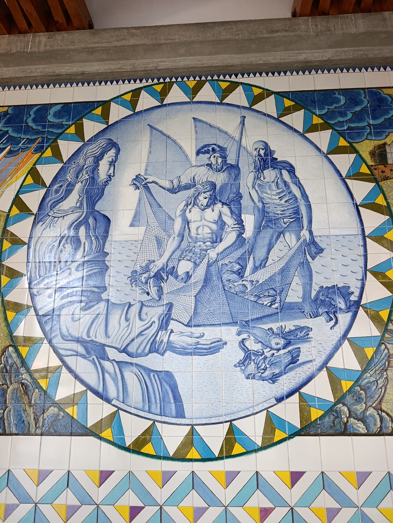 This is a tiled mural in Terras de Bouro, Portugal, depicting a scene of men fishing. The central circular motif showcases three men in a boat, hauling in a net filled with fish. One man, with a beard and wearing a cape, stands in the background. Another man, muscular and bare-chested, is actively pulling the net. A third man, wearing a cap, is also involved in the fishing activity. To the left, a figure with long, flowing hair, possibly a representation of Jesus, extends a hand, seemingly directing the fishermen. The water is depicted with waves, and various fish are scattered around the boat and in the net. The mural is bordered by a decorative pattern of triangles in blue, yellow, green, and white. The background outside the circle includes stylized clouds and hints of landscape, with a building or structure visible on the right. The bottom portion of the mural features a repeating geometric pattern of colorful tiles. The style is reminiscent of traditional Portuguese azulejos.