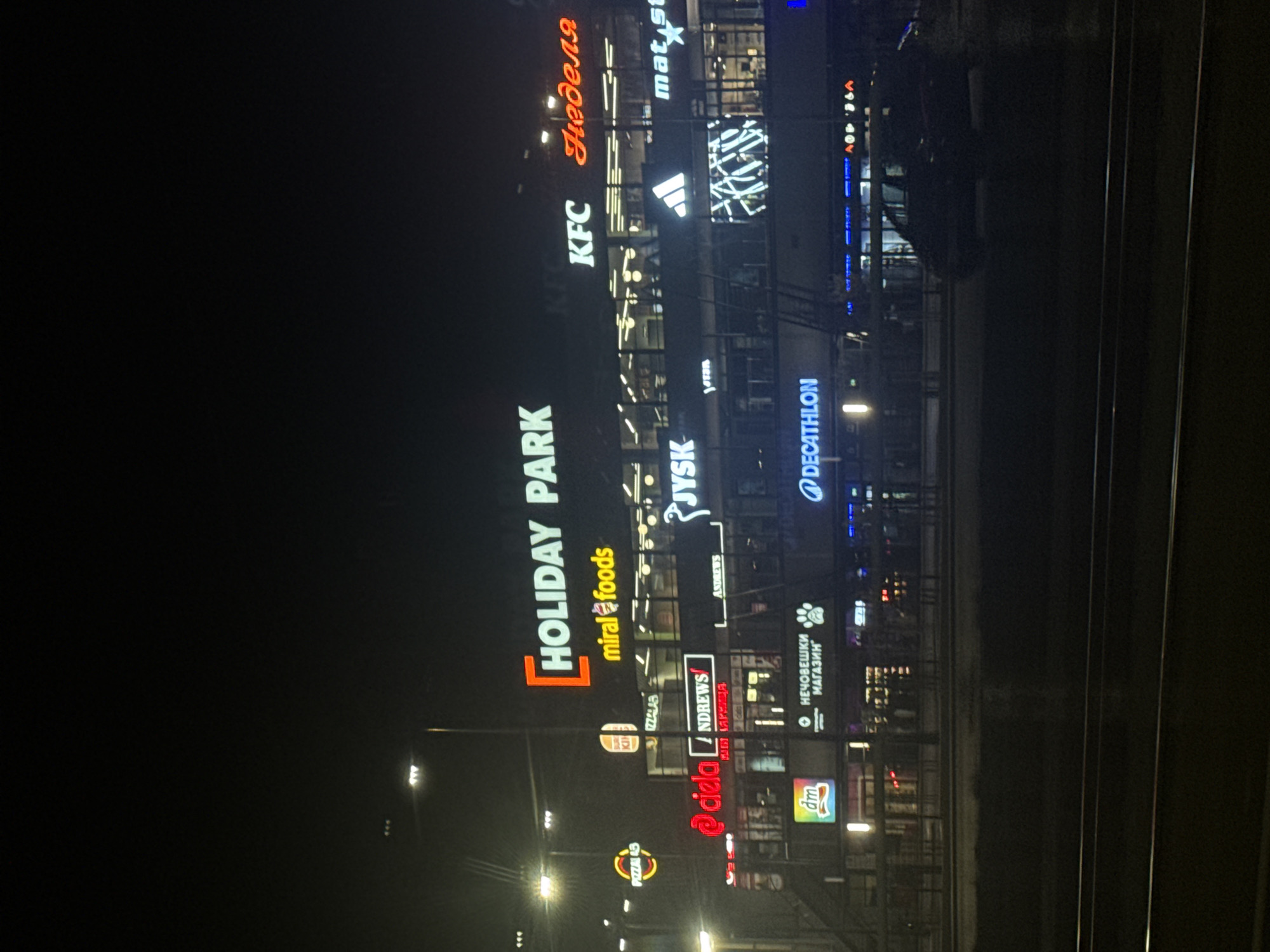 This is a nighttime exterior photograph of a multi-story commercial complex, identified by prominent signage as "HOLIDAY PARK," located in an unknown city, Bulgaria. The building displays numerous illuminated signs for various businesses, including KFC, miral foods, JYSK, Adidas, Decathlon, dm, Burger King (partial), Неделя (Nedelya supermarket), Нечовешки магазин (pet store with a paw print logo), PIZZA 43, ciela, ANDREWS, and mat.star. The sky is dark, indicating night, with artificial illumination from the store signs and several streetlights. In the foreground, a dark street or road is visible, featuring two sets of parallel tram tracks. A dark-colored vehicle, possibly a minivan, is parked or driving on the road in the lower right portion of the image. No individuals are distinctly visible, and no specific actions or interactions are discernible.