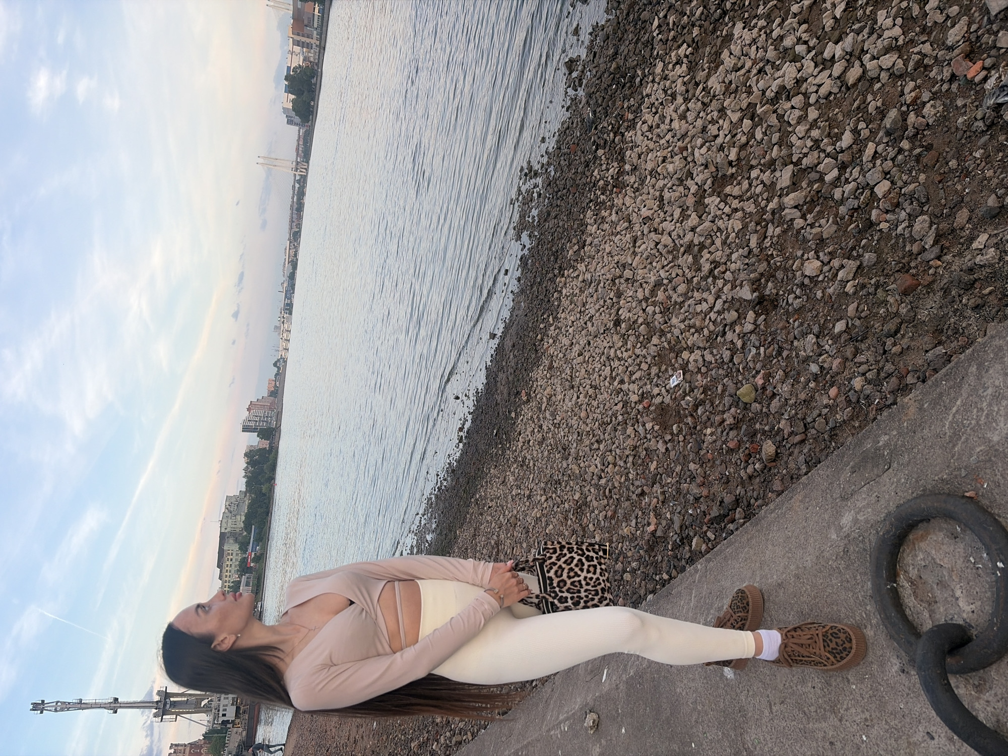A young woman stands on a concrete embankment overlooking a wide body of water in an urban setting, identified as Unknown, Russia. She is positioned facing left, looking towards the distant horizon. Her long, dark hair flows down her back and over her left shoulder. She wears a light beige long-sleeved crop top with a wrap-around design, revealing a black bra and a sliver of her midriff, along with cream-colored ribbed leggings. Her footwear consists of white ankle socks and brown platform shoes with a leopard print pattern. She holds a small, structured handbag with a black and brown leopard print.
To her right, the concrete transitions into a rocky and pebbled shoreline that slopes into the water. The water itself is calm with gentle ripples. In the background, from left to right, a tall industrial crane or tower rises above a dark, ornate building near the shore. Further along the distant bank, a cityscape with various buildings and greenery is visible, culminating in the distinct silhouette of two cable-stayed bridges spanning the water. The sky is partly cloudy with light blue and grey tones, and a faint contrail is present in the upper left. A large, dark metal mooring ring is visible on the concrete in the bottom left foreground.