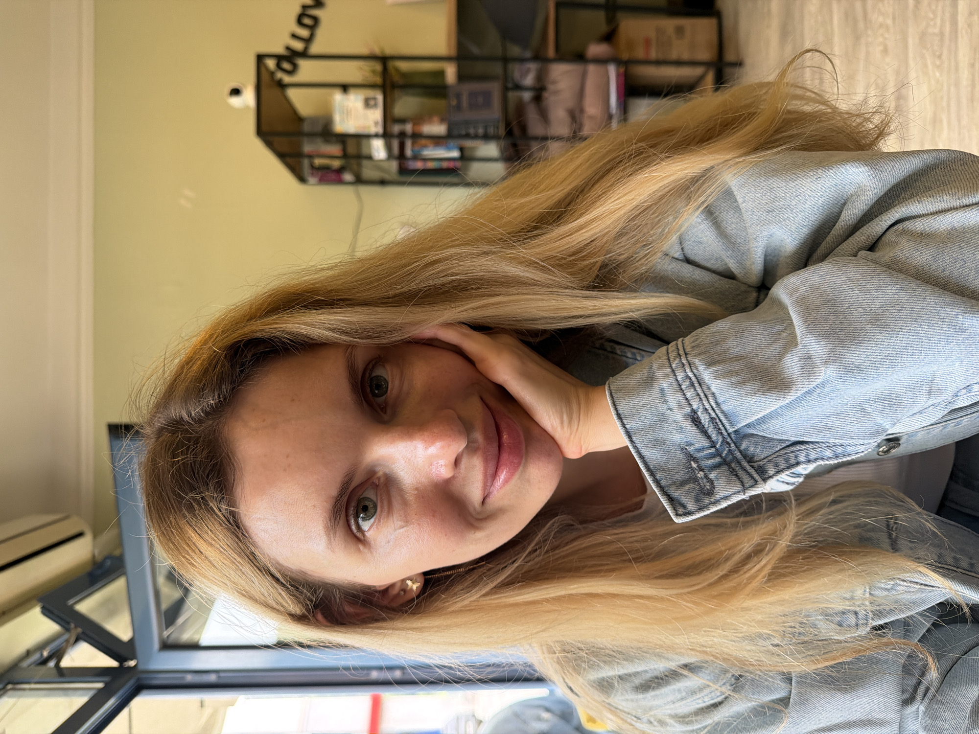 A woman with long blonde hair and a light blue denim jacket is posing for a photo. Her left hand is raised to her cheek. The location is indoors, with a black metal shelving unit visible in the background. Details suggesting the location might be Budva, Montenegro are not discernible from the image.