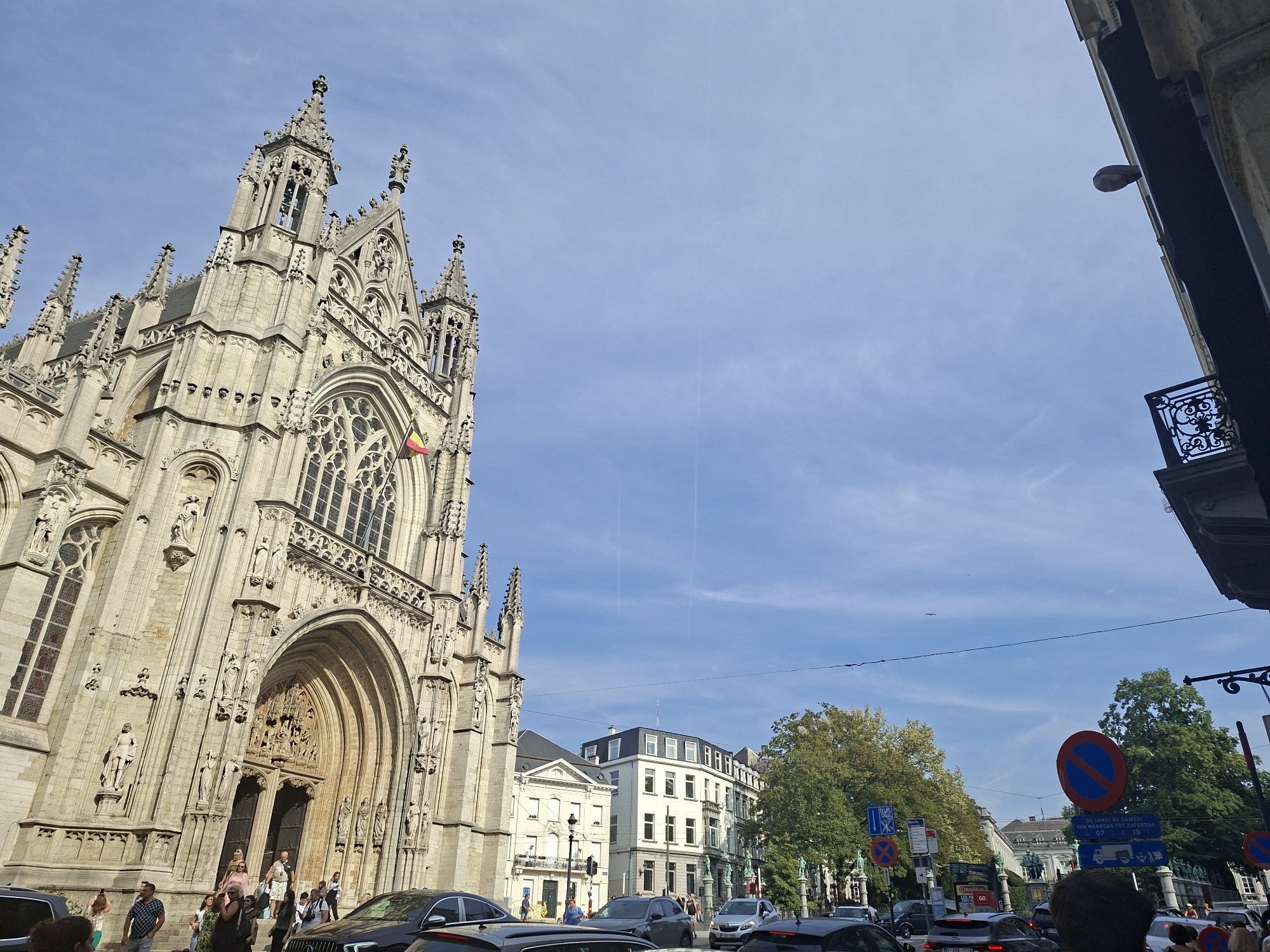 This is an exterior, eye-level, wide-angle shot of the Cathedral of Saint Michael and Saint Gudula in Brussels, Belgium, taken on a clear, sunny day with scattered clouds. The grand Gothic cathedral dominates the left side of the frame, its intricate stone facade and spires reaching towards the sky. A Belgian flag is visible on the right side of the main entrance arch.
In front of the cathedral, several people are seen walking up the steps to the entrance or standing on the sidewalk. The street in front of the cathedral is busy with traffic, including several cars and a bus. Pedestrians are also visible on the sidewalk to the right of the cathedral.
The background features a row of multi-story buildings, some with modern architectural elements and others with more traditional facades. Lush green trees line the street, adding to the urban landscape. Traffic signs, including a no-parking sign, are visible on the right side of the image. The overall scene captures a lively urban environment with a prominent historical landmark.