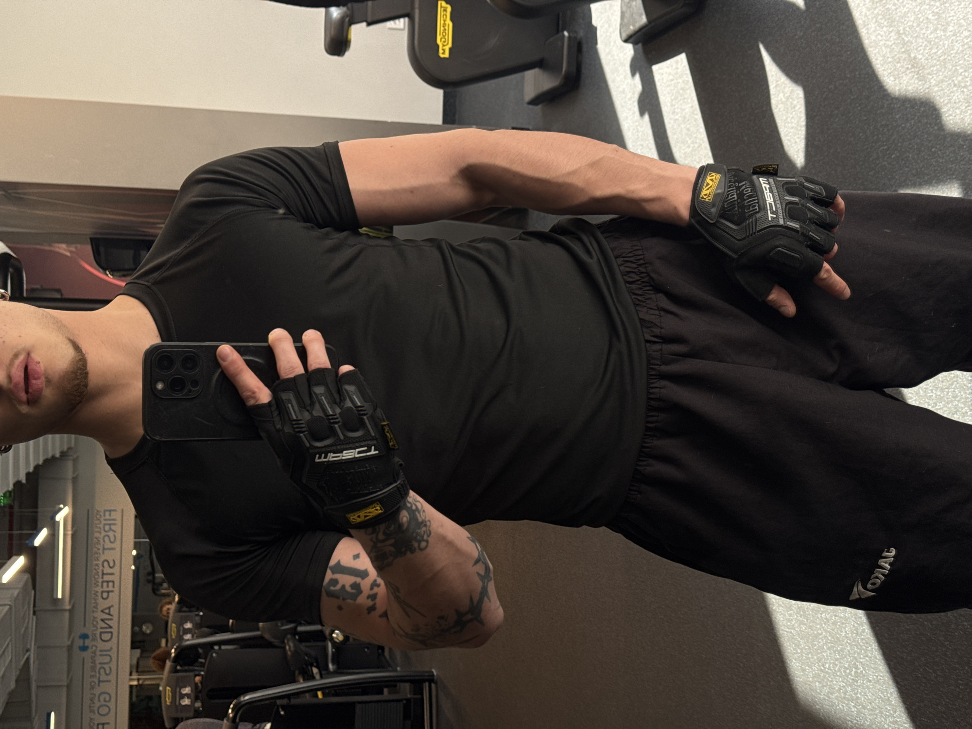 A male individual captures a mirror selfie inside a gym located in Sofia, Bulgaria. The subject is wearing a black short-sleeved athletic shirt, black athletic shorts with a grey "VOYAG" logo (reversed due to reflection) on the left thigh, and black fingerless Mechanix Wear gym gloves. A silver ear gauge is visible in his left ear. His left arm displays multiple tattoos, including the number "13" on the bicep, intricate designs on the forearm, and a barbed wire-like motif around the elbow. Both forearms show prominent veins. A black smartphone with a triple-lens camera system is held in his right hand, partially obscuring his face, which features a neatly trimmed goatee and full lips. The gym environment consists of grey flooring, light-colored walls, and a ceiling with multiple linear light fixtures. In the reflected background, gym equipment such as Technogym exercise bikes and weight machines are visible, along with text on a mirror or wall that reads, "FIRST STEP AND JUST GO / YOU'LL NEVER KNOW WHAT YOU'RE CAPABLE OF." Geometric shadows from sunlight are cast across the floor.