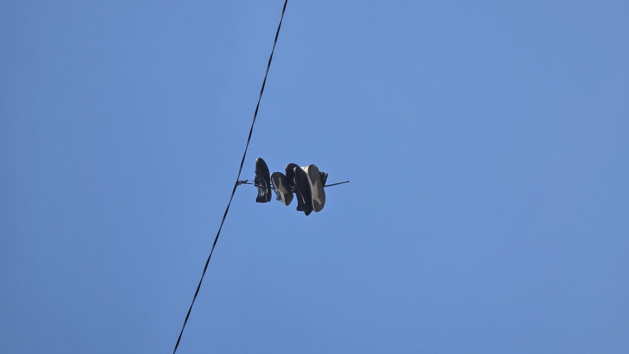 The image shows a pair of shoes hanging on a power line against a clear blue sky. The shoes are mostly dark in color, but there are a couple of light-colored pairs. The picture shows a single power line running diagonally across the frame, with the shoes clustered around one point on the wire. The location of the image is Blagoevgrad, Bulgaria.
