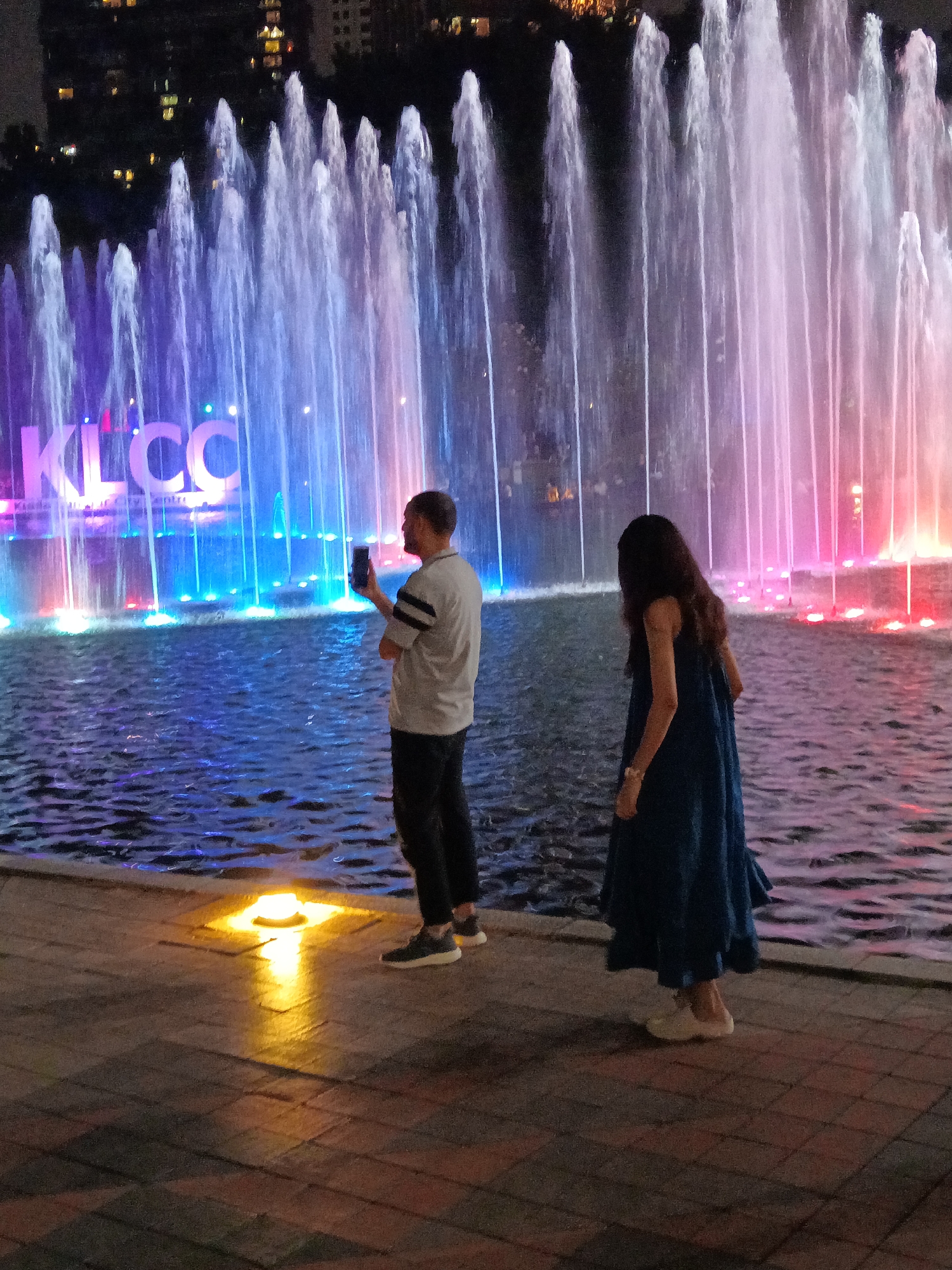 The image shows a view of the Symphony Lake at the base of the Petronas Twin Towers in Kuala Lumpur, Malaysia. Two people are standing by the edge of the lake, which is illuminated by colorful lights and surrounded by a choreographed fountain display. In the foreground, a man in a grey shirt and dark pants is holding up his phone, appearing to take a picture or video of the fountains. To his right, a woman in a dark blue dress is facing away from the camera, also looking towards the fountain show. The Petronas Twin Towers are visible in the background, with the "KLCC" lettering illuminated on a structure behind the fountains. The scene is set at night, with the lights of the city and the fountain display creating a vibrant atmosphere.