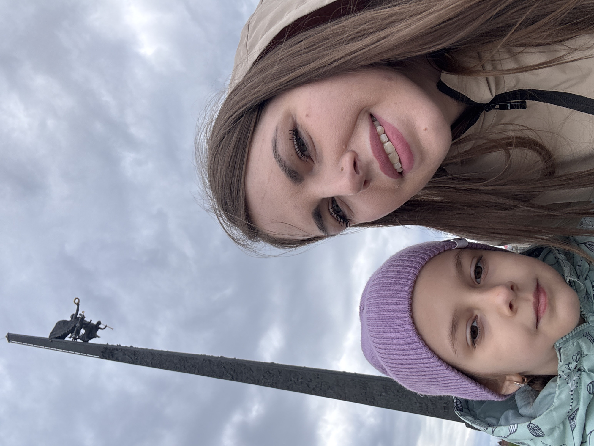 The image depicts a woman and a young girl in Moscow, Russia, taking a selfie with the Monument to the Conquerors of Space in the background. The woman, wearing a beige jacket, is smiling at the camera. The girl is in the bottom right, wearing a lavender beanie. The Monument to the Conquerors of Space is a tall, grey obelisk with a statue at its top, set against a cloudy sky.
