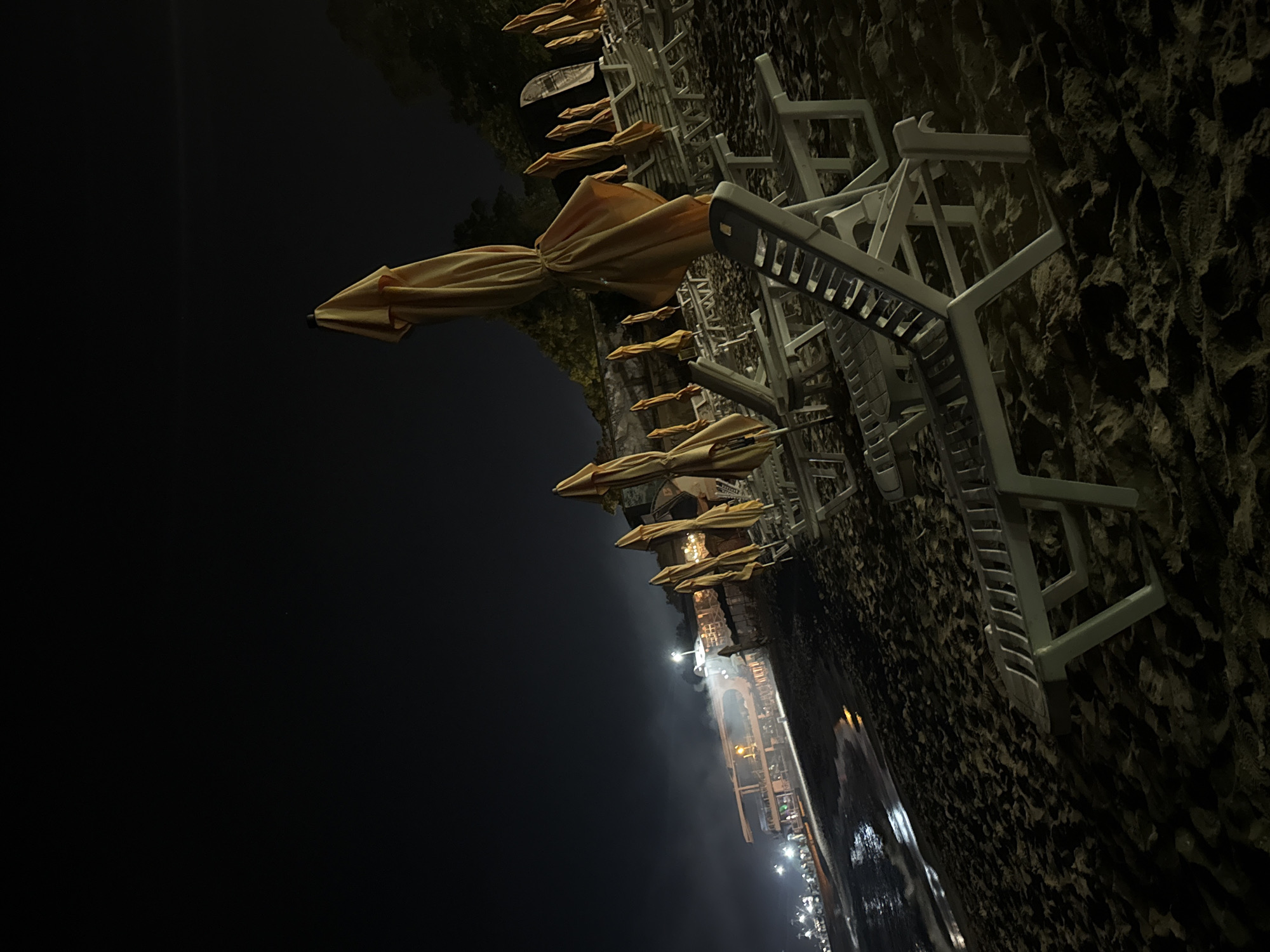 The image depicts a nighttime scene on a sandy beach in Varna, Bulgaria. In the foreground and midground, numerous white plastic beach lounge chairs are arranged in rows, with many yellow-tan, folded beach umbrellas standing upright beside them. The sand shows various textures and shadows, with faint imprints of what appear to be footprints. To the left, the dark water of the Black Sea is visible, with illuminated ripples and a thin line of white foam where gentle waves meet the shore. Further along the coastline to the left, a multi-level building or complex is illuminated by artificial lights, with a noticeable plume of light-colored mist or smoke rising from its vicinity. Beyond this structure, additional lights stretch along the distant coastline, indicating a populated urban area. The background consists of a dark night sky, with a dark line of foliage or trees visible behind the beach furniture on the right. No individuals are present in the scene, which appears deserted. The primary light sources are artificial lights from the distant structures and along the shore, creating high contrast and deep shadows across the beach.