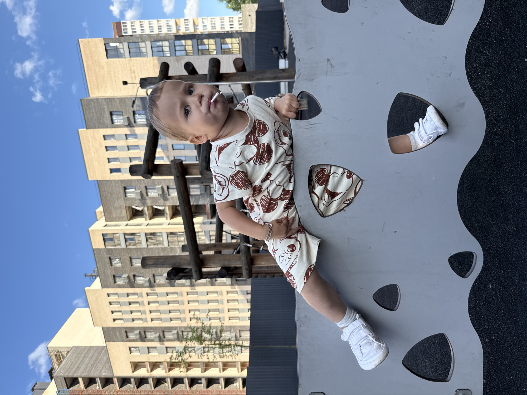 A young Caucasian child with blond hair and blue eyes is playing on a playground in Rusanovo, Russia. The child is wearing a beige outfit with a brown Mickey Mouse pattern and white sneakers. They are holding a lollipop in their mouth and are leaning over a grey wall with cutouts. In the background, there are modern apartment buildings under a bright blue sky with scattered clouds. The playground surface is dark, textured material.