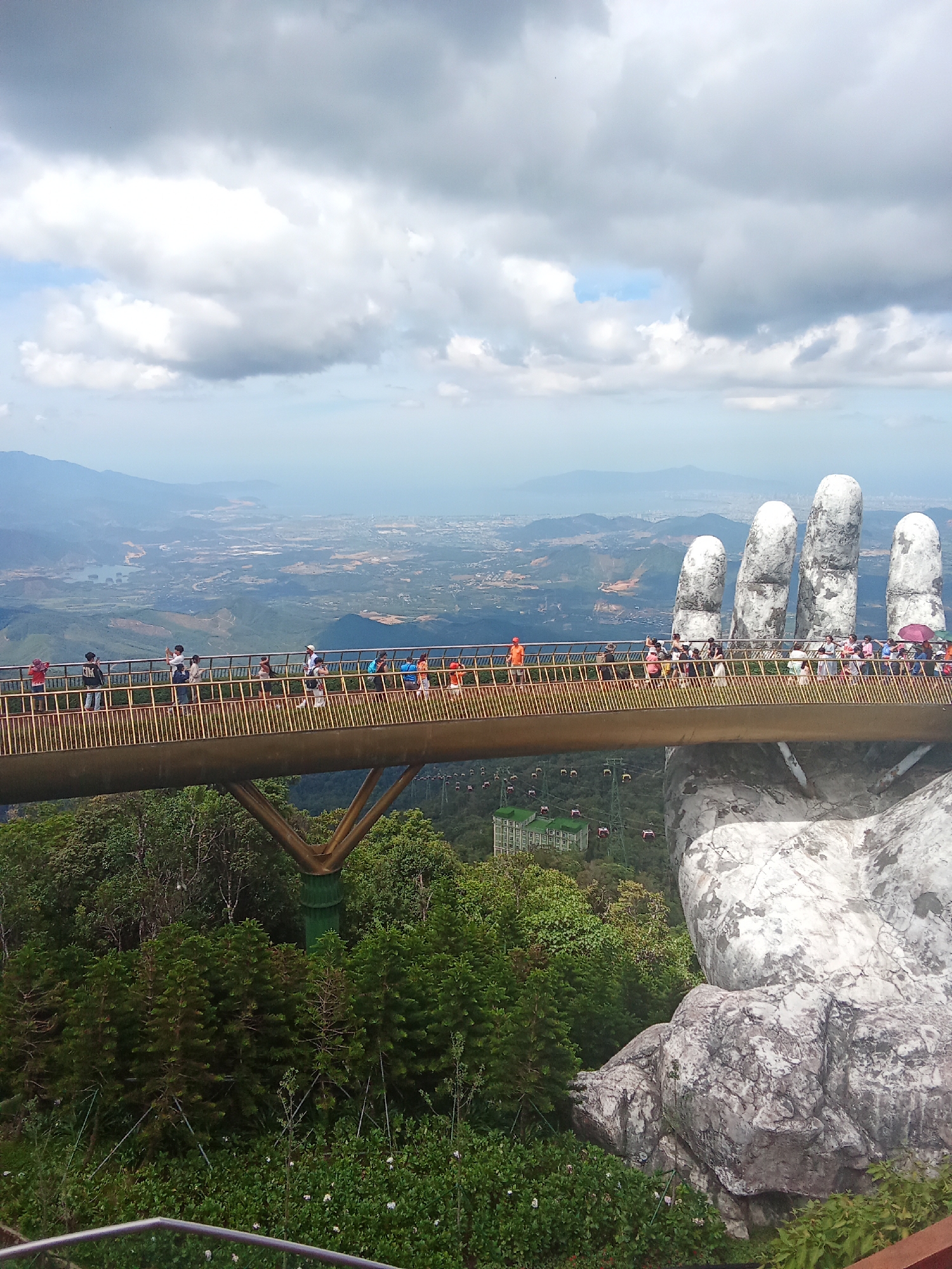 A wide shot captures the Golden Bridge in Da Nang, Vietnam, an architectural marvel designed as two massive hands emerging from the mountainside supporting a pedestrian walkway. The bridge is adorned with a golden railing and populated by a diverse group of people walking and taking photographs, all bathed in the soft light of an overcast sky. Below the bridge, a lush green forest stretches out, punctuated by a series of cable cars descending into the valley. In the distance, the cityscape of Da Nang is visible, nestled between rolling hills and the vast expanse of the ocean. The scene evokes a sense of awe and wonder, a testament to human ingenuity and the breathtaking beauty of the natural landscape.