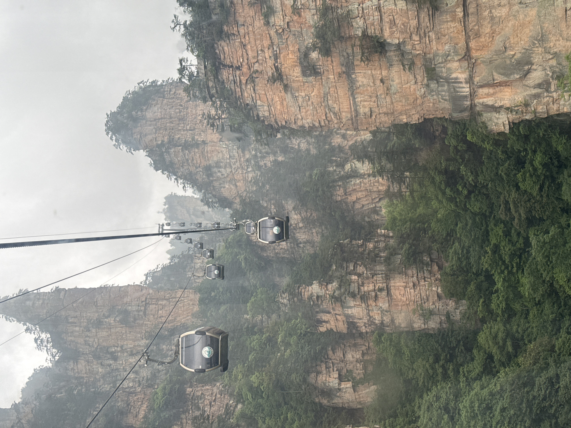 The image shows a series of gondola cars suspended on cables in a mountainous landscape. The gondolas are grey and black with a circular logo on the side, and they are oriented diagonally across the frame. The background features towering rock formations with vertical striations, covered in patches of green vegetation. A thick haze or fog obscures parts of the landscape, adding to the sense of depth and atmosphere. The location is Wulingyuan, China.