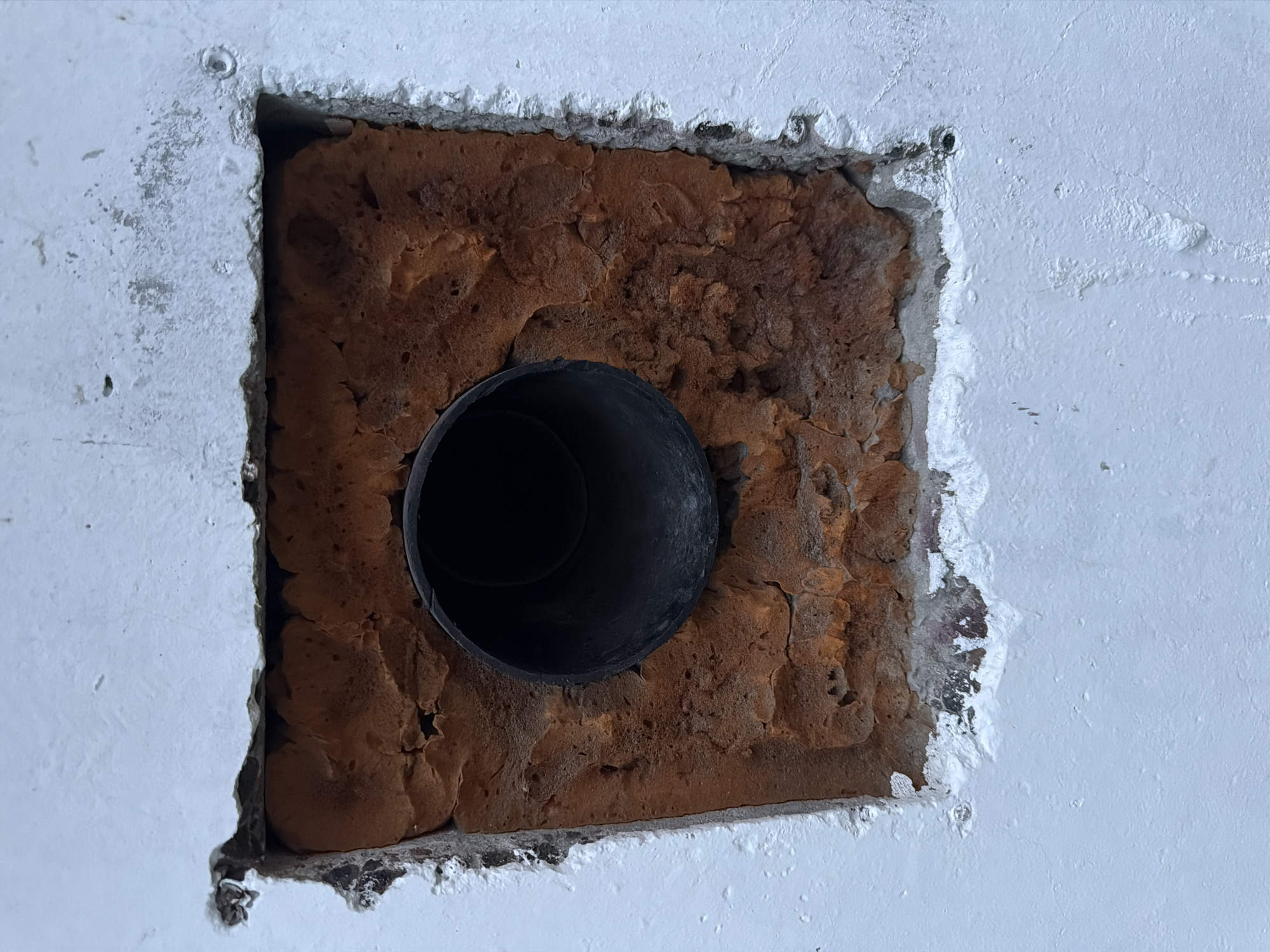 The image shows a close-up, eye-level view of a rough, rectangular opening in a white painted wall. The opening reveals a cavity filled with reddish-brown, porous material, possibly insulation or crumbling brickwork. A black, circular pipe or vent protrudes from the center of this cavity, with its opening facing the viewer. The edges of the opening in the white wall are uneven and chipped, with some white paint flaking away. The texture of the white wall appears somewhat rough and uneven. The lighting is even, suggesting an outdoor or well-lit indoor environment. There are no people, animals, or any other discernible objects or activities visible in the frame. The image does not provide any specific clues to indicate its location within the city of Surgut, Russia, or any other geographical area.