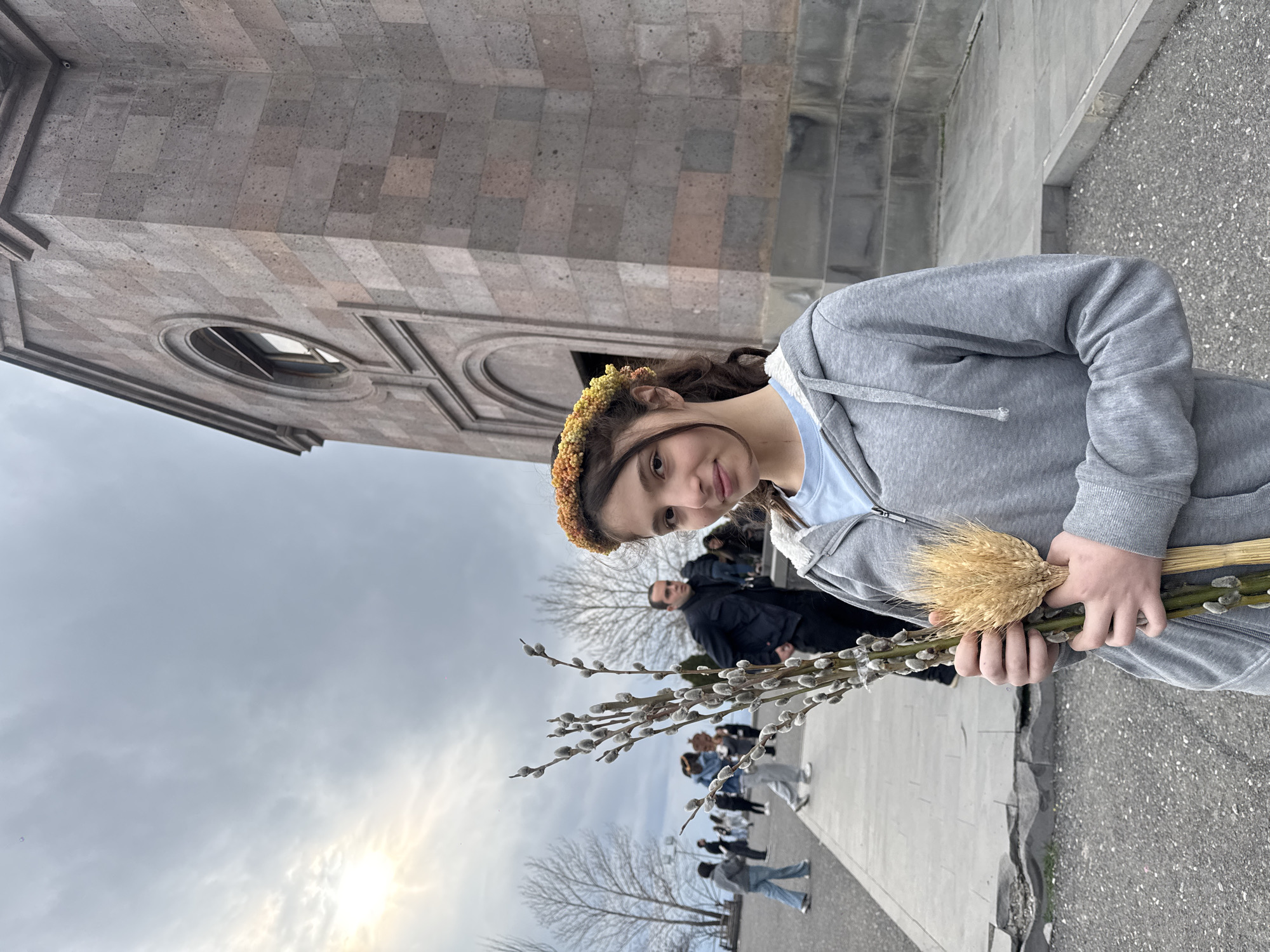 A slightly rotated eye-level shot shows a girl holding wheat and willow branches in front of a stone building in Yerevan, Armenia. She has brown hair, a floral crown, and wears a gray hooded sweatshirt and a blue shirt. People walk on a sidewalk behind her, and leafless trees are visible in the background. The cloudy sky suggests overcast weather.