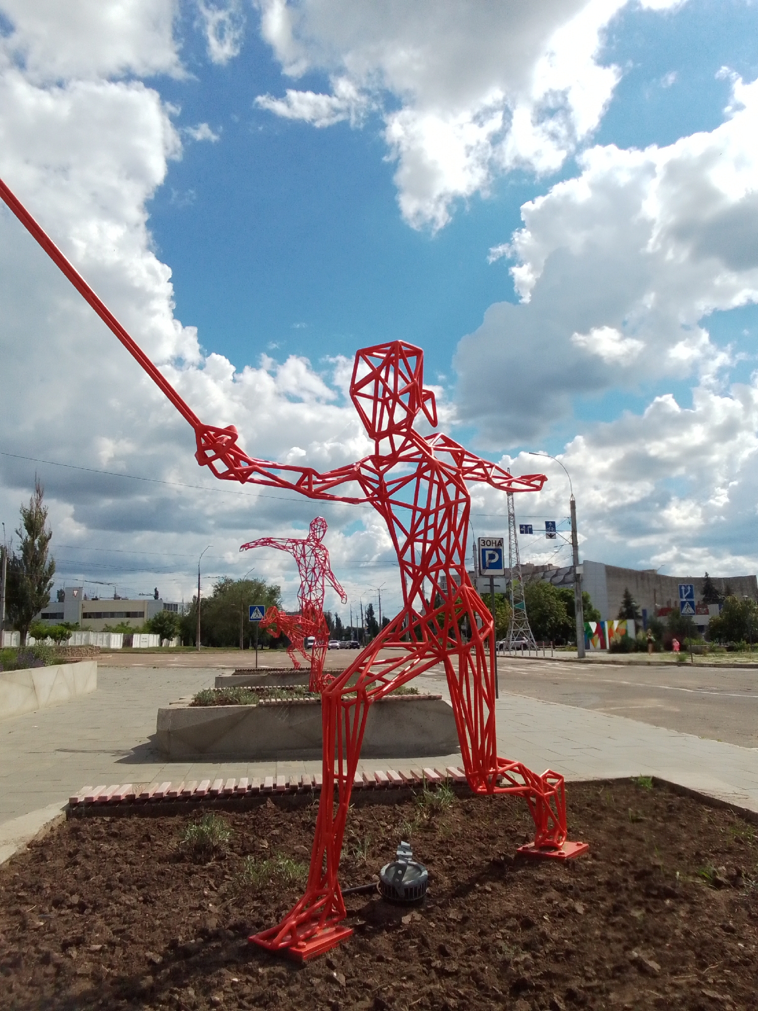 A tall, bright red, wireframe sculpture of a fencer is the central focus of this outdoor, urban scene. The sculpture is positioned in a planter filled with dark soil and some sparse greenery, with a concrete walkway and planter boxes in the background. The fencer sculpture is in mid-action, with one arm extended holding a long, thin red rod resembling a saber, and the other arm bent. Its legs are in a lunging pose. A smaller, similarly styled red wireframe sculpture of another fencer is visible behind and to the left of the main sculpture, also in a dynamic pose. In the background, a street with traffic lights, road signs, and buildings can be seen under a bright blue sky with fluffy white clouds. A solitary thin, tall green tree stands to the left of the frame.