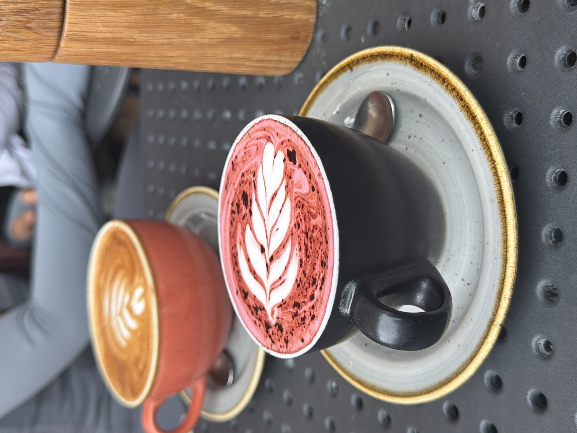 The image shows two cups of latte art coffee on a grey, perforated metal table. One cup is black with reddish-pink latte art resembling a leaf, and the other is reddish-orange with brown latte art. Both cups sit on saucers and have spoons. A light-colored wooden surface is visible at the top of the frame. A person in a grey outfit is also partially visible in the background. The location is presumed to be a café in London, United Kingdom, based on the coffee culture.