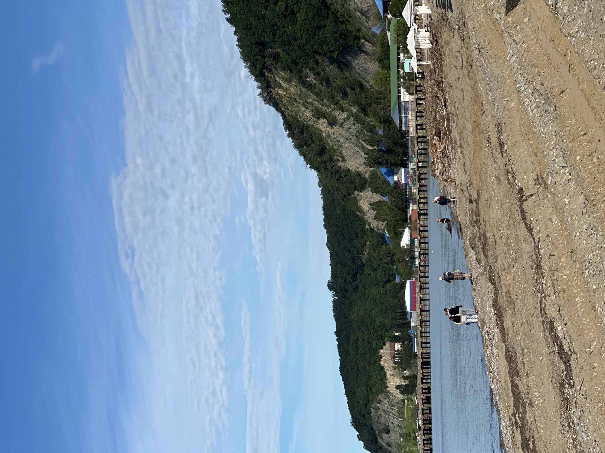 The image captures a coastal landscape in Lermontovo, Russia, under a clear blue sky with scattered white clouds. A calm, blue body of water, likely the sea, occupies the left side of the frame. To its right, a sandy and pebbly beach stretches, showing some tire tracks or indentations and a small amount of driftwood near the water's edge.
In the background, a steep, green hillside or mountain range, densely covered with trees and patches of exposed rock, rises dramatically, separating the sky from the coastline. Along the base of this hillside, bordering the water and the beach, several buildings are visible. These include structures with white walls and roofs, some featuring blue or green roofing, indicative of a resort or settlement. White beach umbrellas are also present near some of these buildings. A pier-like structure with vertical posts extends along the water's edge, running parallel to the beach and buildings.
Several people are depicted on the beach or wading in the shallow water. In the foreground, two individuals stand near the water's edge; one wears a dark top and light pants, and another is partially obscured. Further along the shore, another person, wearing a dark top and a hat, is visible near the water. Additional figures are seen in the distance, leisurely enjoying the coastal environment.
The scene suggests a bright, sunny day, likely late morning or afternoon, with pleasant weather. The overall atmosphere is one of tranquility and relaxation. There is no visible text in the image.