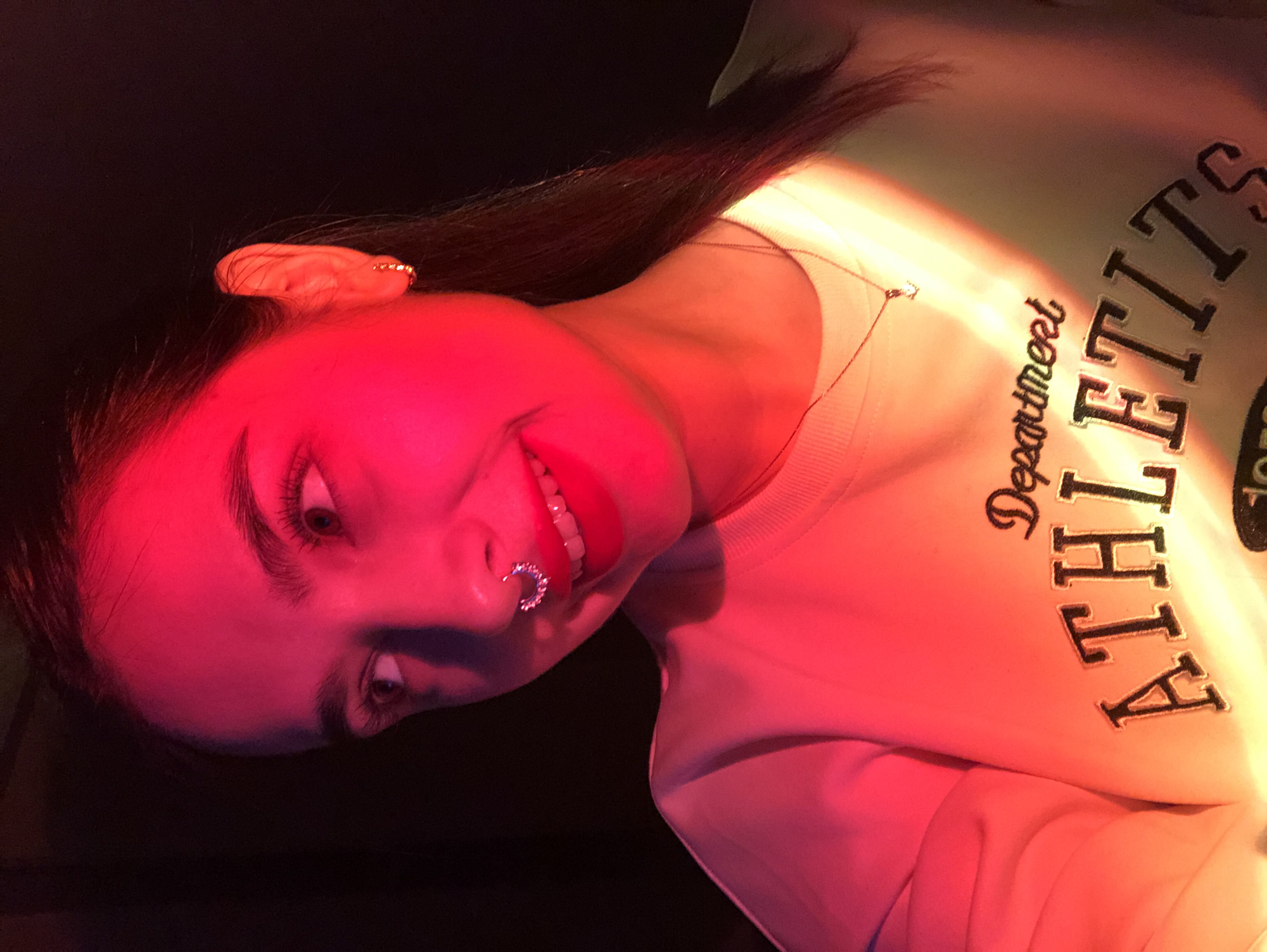 This media file is a tilted, close-up portrait of a young woman, illuminated by dramatic red and orange artificial lighting. Her face, dominating the left side of the frame, is strongly lit by a vibrant red hue, while her upper body and clothing on the right are bathed in a warmer, softer orange-pink glow.
The woman has dark hair, styled to fall behind her shoulders, and distinct dark eyebrows. She wears red lipstick and has a sparkling, multi-studded septum piercing. Her expression is playful, characterized by a wide smile revealing her teeth, and her eyes, enhanced with eyeliner and mascara, look slightly upwards and to the side. A small, golden earring is visible on her left earlobe.
She is wearing a light-colored, possibly cream or off-white, long-sleeved sweatshirt. A delicate, thin chain necklace with a small pendant rests on her neck. Visible text on her sweatshirt includes "Department" in a script font and partially "ATHLETITS" in large, bold, block letters, likely part of "ATHLETICS." The background is dark and indistinct, focusing attention on the subject and the striking lighting. The scene appears to be indoors, possibly an evening setting, in a location like Noviy, Russia, though specific cues for Russia are not visible in the image itself. The overall mood is casual and slightly enigmatic due to the intense lighting.