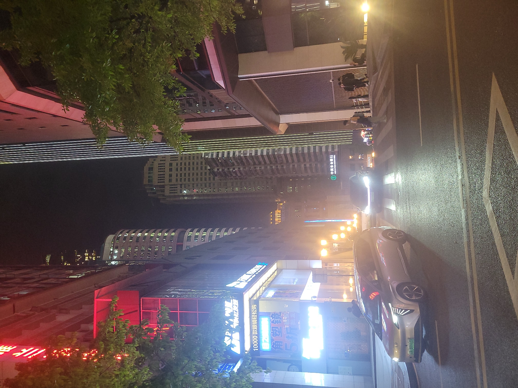The image captures a vibrant night scene in Yuzhong District, China, showcasing a bustling urban environment illuminated by numerous artificial lights. The overall setting is a city street flanked by modern, tall buildings.
In the foreground, a wet asphalt road stretches diagonally across the frame, marked with bright yellow double lines, a broken white line, and a pedestrian crosswalk. A sleek, light-colored sedan with its headlights on is visible on the right side of the road, facing away from the viewer. Several people are seen walking on or near the crosswalk and sidewalks, some appearing to cross the street.
The background is dominated by an array of buildings. On the left, a building features distinct horizontal lines of white light across its facade. Further back, a unique structure with a curved, almost cylindrical top and numerous illuminated windows stands out against the dark sky. Other buildings feature illuminated signs and windows, creating a glow that reflects off the wet street surface. Lush green foliage from trees is visible in the upper left and lower right corners, adding a natural element to the urban landscape.
The time of day is clearly night, with the sky completely dark. The ground appears wet, reflecting the intense artificial lighting from streetlights, building facades, and numerous neon signs. The strong illumination creates a dynamic and energetic atmosphere.
Visible text on various neon signs includes Chinese characters. One prominent blue sign displays "10000 贷款轻松购" (10000 loan easy purchase), while a green sign with a white logo reads "中国邮政储蓄银行" (China Post Savings Bank). Other glowing red and blue signs display further Chinese text, some of which are partially obscured or difficult to decipher due to brightness.