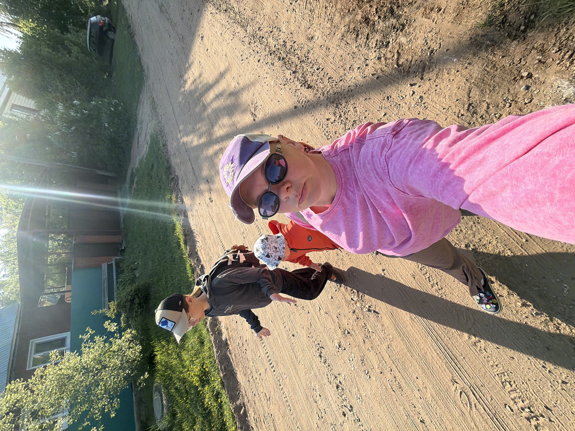 Three individuals stand on a dirt road on a sunny day. The person nearest the camera is an adult wearing a pink shirt, tan pants, a purple cap, and sunglasses. Two children stand adjacent to them. One is wearing a gray hoodie and a baseball cap, and the other is wearing an orange hoodie and a patterned bucket hat. The background contains a house with green trim and a silver car parked in a grassy area. The location is Chaikovsky, Russia.