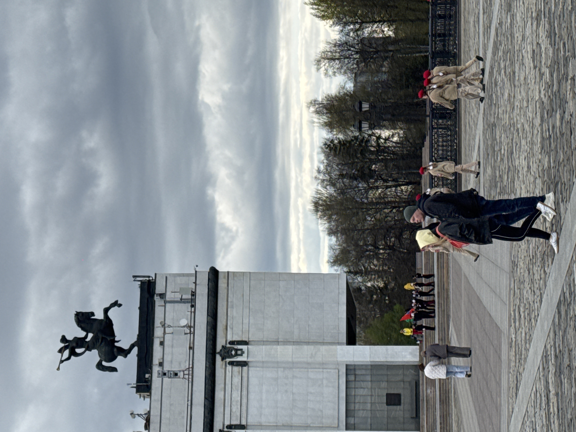 The image shows the Victory Monument on Poklonnaya Hill in Moscow, Russia. The monument features a tall, rectangular structure with a statue of Saint George slaying the dragon at its base. A group of people, some wearing red berets and uniforms, are visible in the background. Other people are walking along the paved area near the monument. The sky is cloudy and overcast. Trees line the area behind the paved space. A railing is visible in the middle ground.