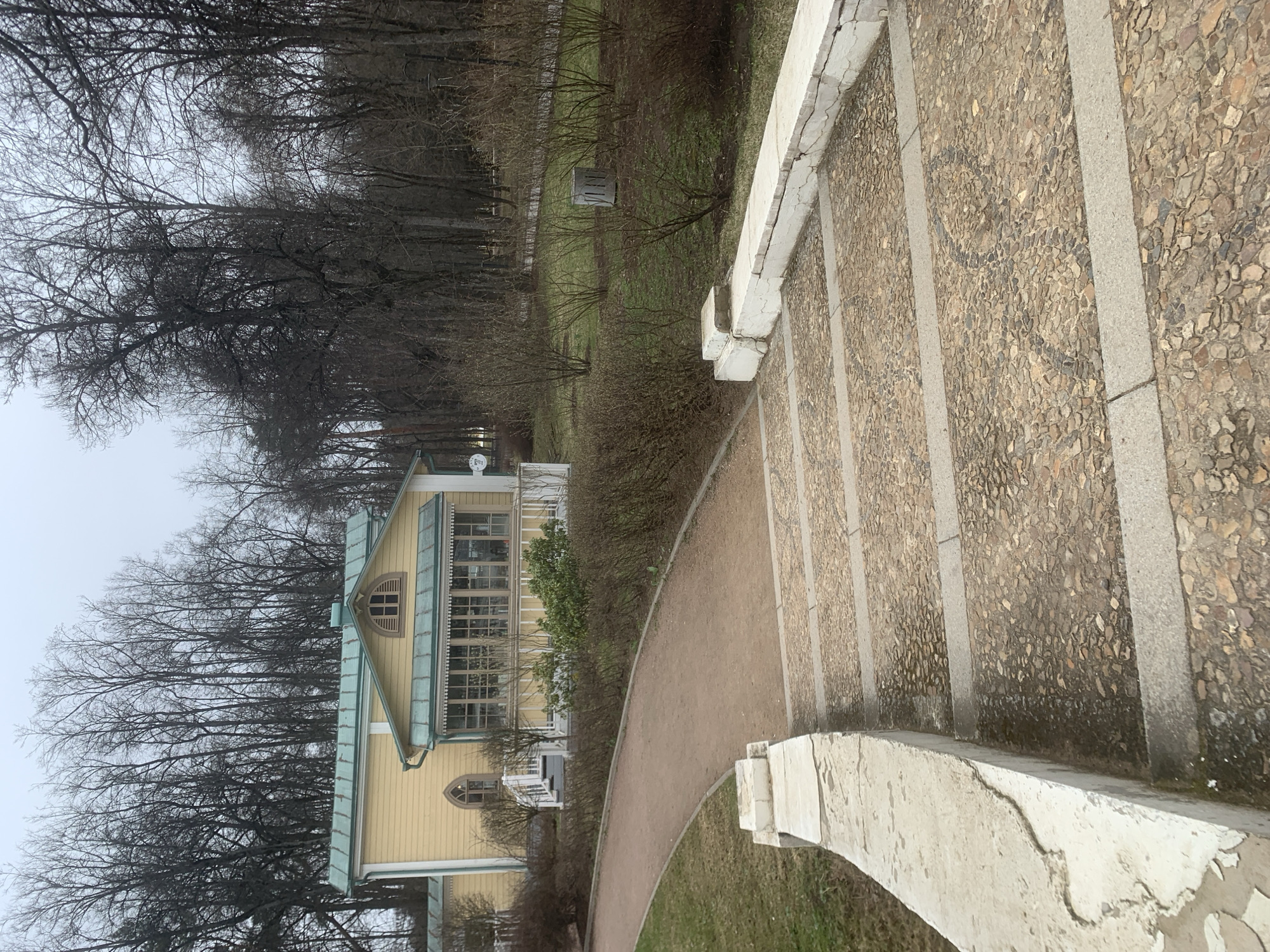 The image depicts a building with pale yellow siding and a pale green roof, located in a park setting. Bare trees are prominent in the background against an overcast sky. A path leads from the building towards the foreground, where there are stone steps with a white stone railing. The stone steps are made of small rocks cemented together. The location is likely in the city of Moscow, Russia.