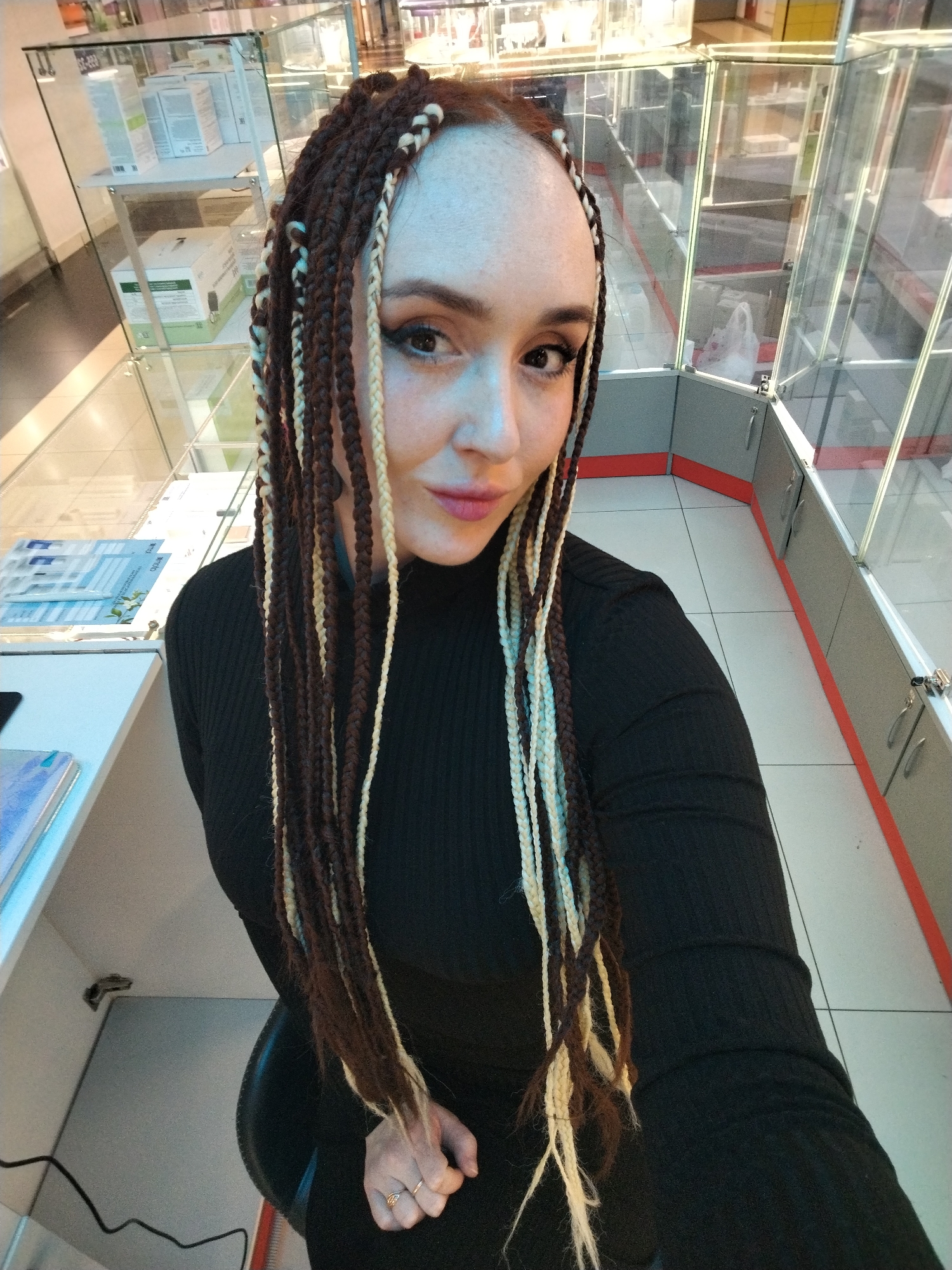 A selfie-style photograph shows a woman with light skin and red hair partially visible at the scalp, styled in brown and blonde braids. She is wearing a black long-sleeved turtleneck top. The woman is seated in a chair and facing the camera.
The background consists of glass display cases, white tile flooring with a red border along the base of the cases, and an adjacent counter area with brochures or pamphlets on it.
The setting appears to be a commercial space, possibly a store in Izhevsk, Russia.