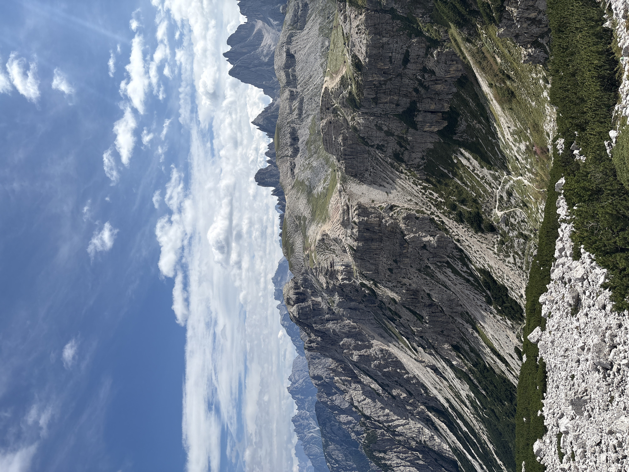 The image shows a panoramic view of the Tre Cime di Lavaredo, a mountain range located in the Dolomites, within the municipality of Auronzo di Cadore, Italy. The foreground is dominated by a rocky, scree-covered slope with scattered evergreen shrubs. A winding path is visible descending into a valley dotted with more shrubs and rocks.
The midground features a dramatic, rugged mountain face with steep cliffs and rocky outcrops. Vegetation, primarily grasses and shrubs, clings to the less steep sections. Another winding path is visible here, with several small figures of people walking on it, suggesting a hiking trail.
In the background, a series of sharp, jagged peaks rise against a bright blue sky filled with scattered white clouds. The overall impression is of a vast, mountainous landscape with a clear, sunny day. The terrain is steep and rocky, typical of alpine environments.