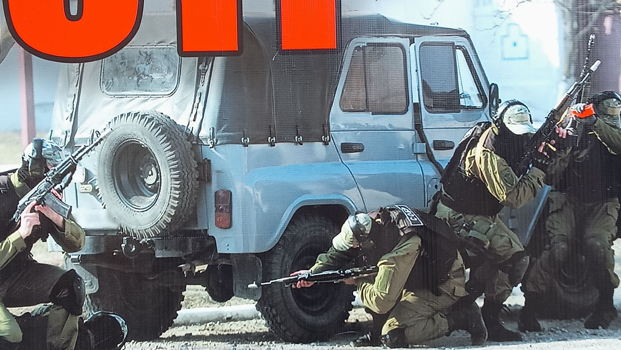 This image depicts an outdoor tactical training exercise or demonstration involving at least five individuals in special operations gear, using a utility vehicle for cover. The scene is set in Tiraspol, Moldova, under clear daytime conditions.
The central subject is a light blue or grey UAZ-452 van, commonly known as a "Bukhanka," featuring a canvas top on its rear section and a spare tire mounted on the back. This vehicle is being utilized by the operatives for concealment and cover during the drill.
The individuals are dressed in olive green tactical uniforms, dark tactical vests (possibly plate carriers), and balaclavas or other head coverings that obscure their faces, with some also wearing baseball caps. They are armed with AK-style assault rifles; some of these rifles have distinct orange safety markers, indicating a training scenario. The operatives are shown in various defensive or attacking stances around the vehicle, aiming their rifles. One individual kneels near the rear wheel, another crouches by the front passenger door, and others are visible to the right, also in ready positions.
Notable details include patches on the backs of at least two individuals' vests, which clearly display the Cyrillic letters "СОБР" (SOBR), standing for *Spetsialny Otryad Bystrogo Reagirovaniya* (Special Rapid Response Unit). In the background, a light-colored wall features a painted target silhouette, further confirming the training nature of the event. Large, bright orange letters from an external sign or banner are partially visible at the top of the frame, partially obscuring the vehicle.
