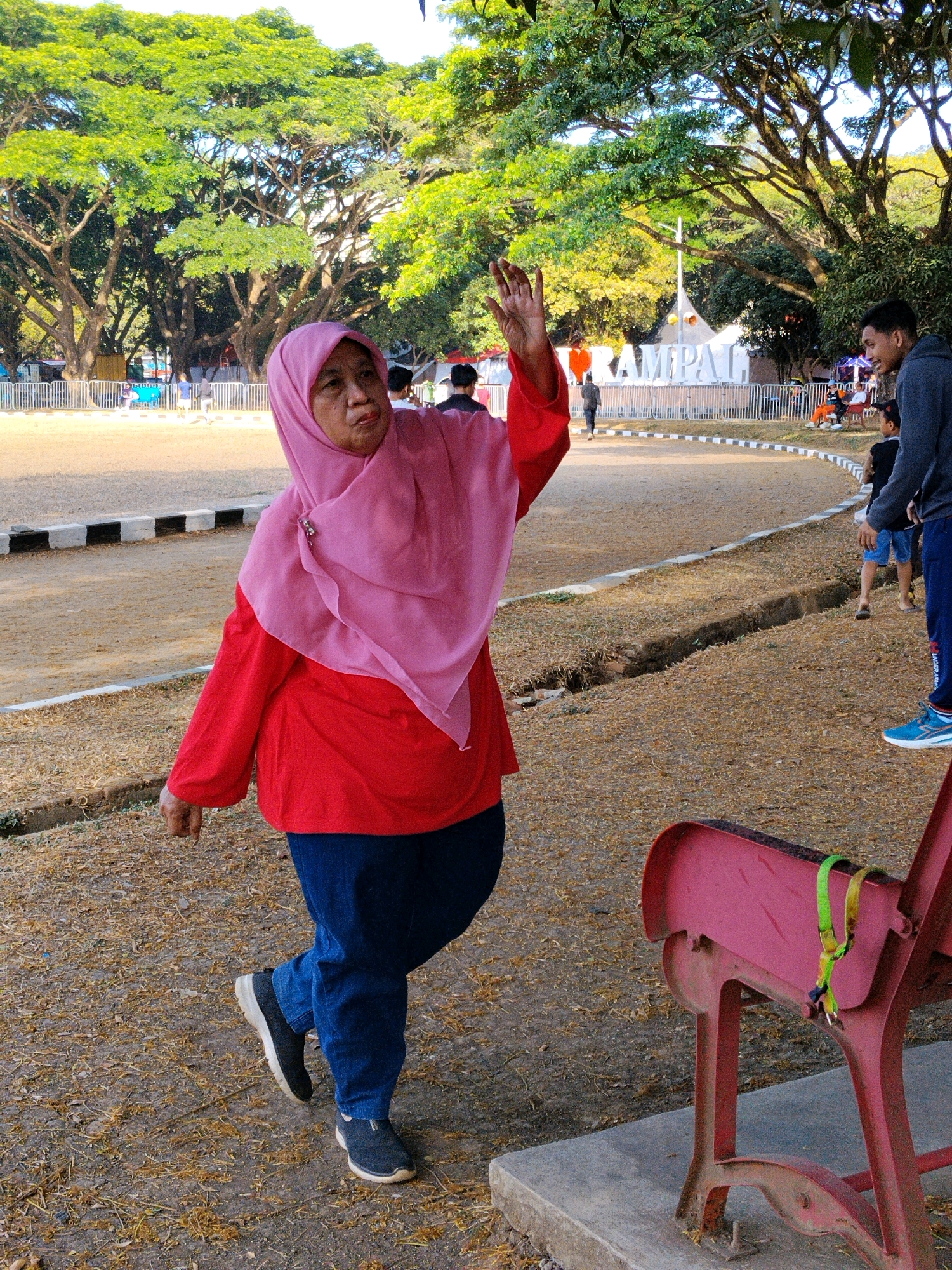 An elderly woman wearing a pink hijab and a red long-sleeved shirt is walking forward and waving her right hand in an outdoor area. She is also wearing dark blue pants and black sneakers. In the background, there are large trees with green leaves, a white fence, and a large sign that reads "RAMPALL". There are other people in the background, including a young man in a gray hoodie and blue pants, and children. The ground is covered with dry grass and dirt. A red bench with a green strap is visible in the foreground. The scene appears to be in a public park.