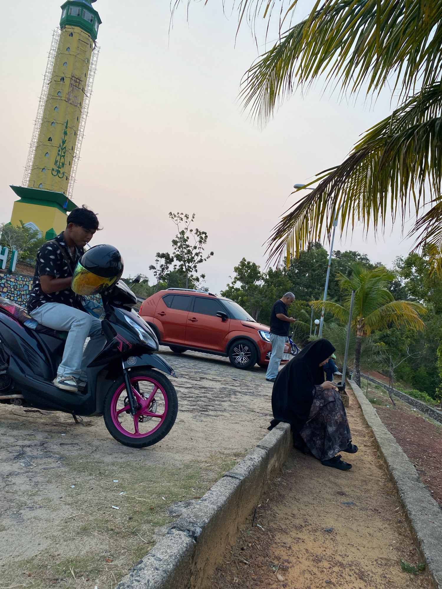 In Tanjung Pinang, Indonesia, on a dirt path near a tall, yellow tower structure with green accents and scaffolding, a young man wearing a black patterned shirt and light blue jeans sits on a black scooter. He holds a black helmet with a yellow and iridescent visor. A bright orange compact car is parked behind the scooter. To the right, two individuals are seated on a concrete curb. One is a woman wearing a black hijab and a patterned dress, looking at her phone. Another person, possibly a man in a dark shirt, stands near the car, also looking at a phone. Palm fronds overhang the scene, and lush green trees and vegetation form the background under a hazy sky.