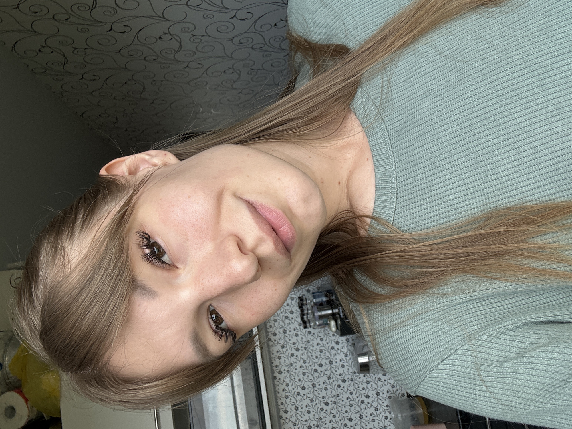 A young woman with light brown hair, wearing a light green ribbed long-sleeved shirt, is looking at the camera and smiling. She has light skin with freckles, brown eyes, and noticeable eyelashes. Her hair is styled with bangs. The background is out of focus and appears to be an indoor setting, possibly a kitchen, with patterned wallpaper visible on the right and some objects on a counter or shelf on the left. The overall lighting suggests it is daytime. The image is a close-up selfie.