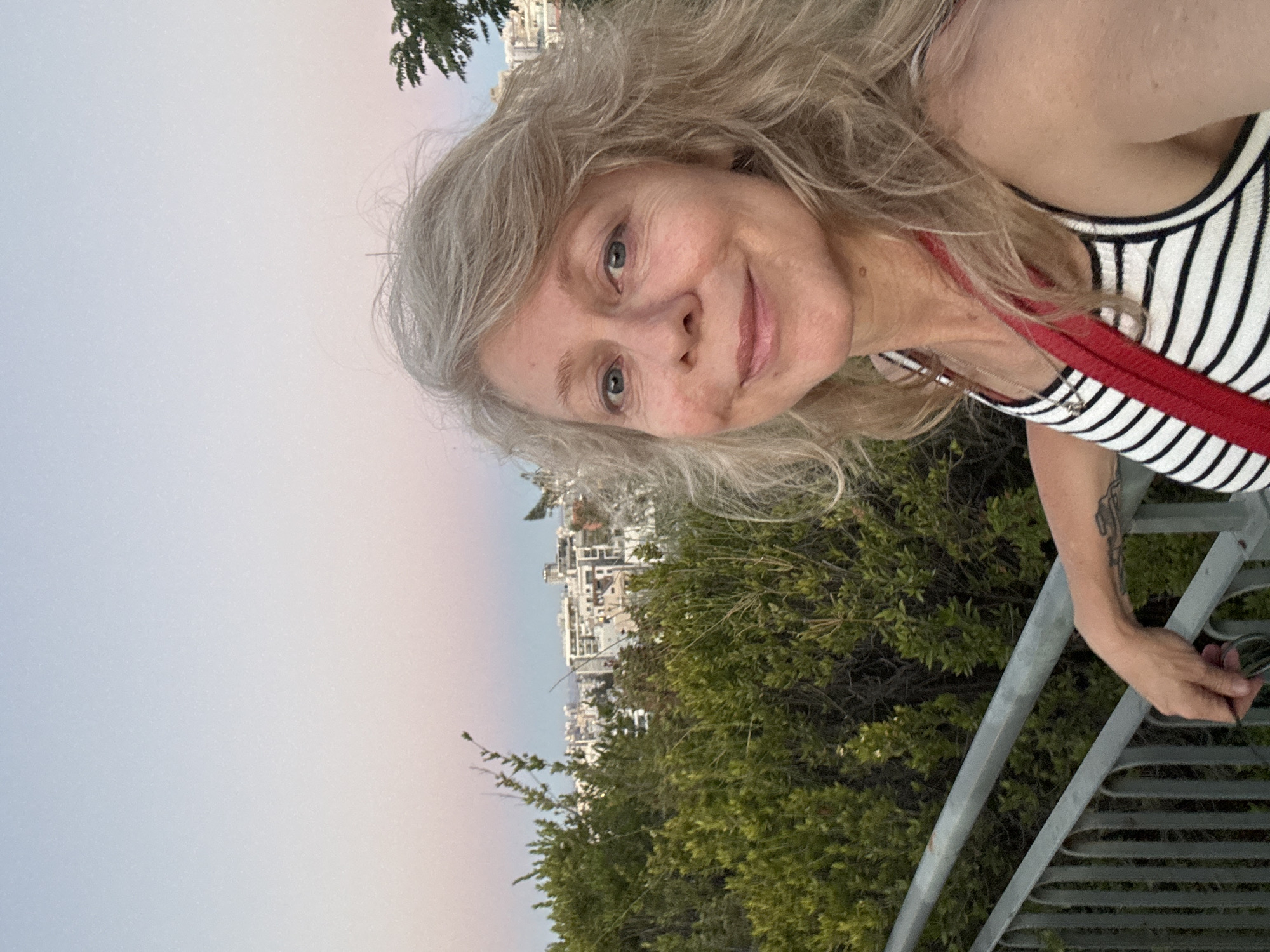A woman with blonde hair is taking a selfie outdoors. She is wearing a striped tank top with a red strap across her chest. Her left arm is resting on a metal railing, and she is holding a pair of glasses in her hand. Behind her, there are green bushes and a city skyline in the distance. The sky is a gradient of pale blue and pink, indicating it is either dawn or dusk. The woman appears to be on a viewpoint overlooking the city of Ramat Gan, Israel, likely from a park or elevated area.