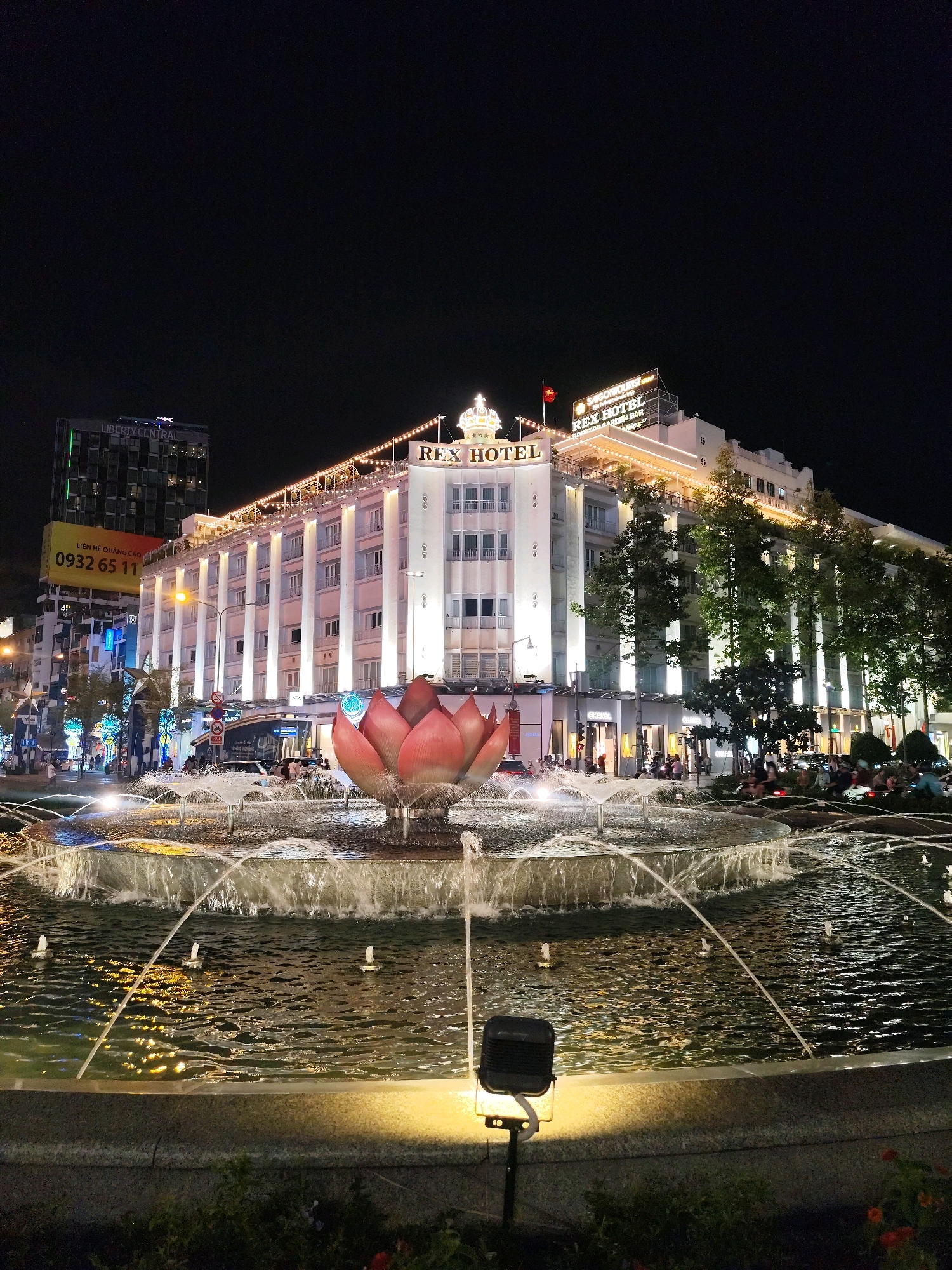 The image captures a nighttime view of the Rex Hotel in Ho Chi Minh City, Vietnam, centered around a large, multi-tiered fountain. The Rex Hotel, an iconic landmark, is illuminated with warm white lights along its facade and rooftop, highlighting its architectural details and a prominent "REX HOTEL" sign adorned with a crown. A Vietnamese flag flies above the hotel. The fountain in the foreground is active, with water jets creating a dynamic display, illuminated by underwater lights and a spotlight on the lower edge of the fountain basin. A large, stylized pink lotus sculpture sits at the center of the fountain. In the background, other buildings, including the Liberty Central hotel, are visible, along with streetlights and illuminated signs. Several people are seated and walking around the fountain area, and a few vehicles are present on the street. The overall scene depicts a vibrant urban environment at night.