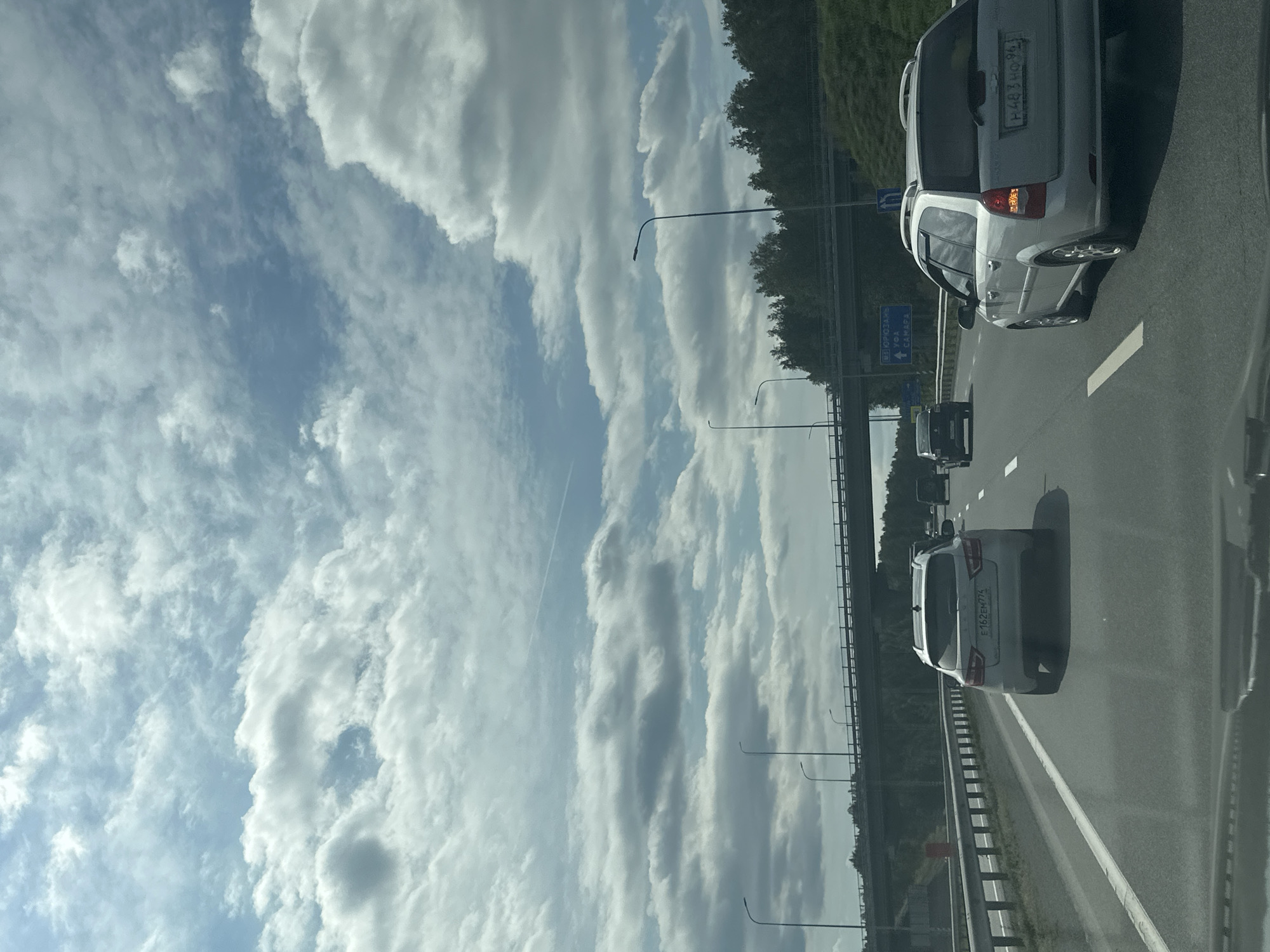 This is a medium shot from inside a vehicle traveling on a multi-lane highway. The sky is filled with a mix of white and gray clouds, with patches of blue visible. A highway overpass with multiple lamp posts is visible in the distance. Trees line the right side of the highway, and a grassy embankment is also present. Several cars are on the highway, including a silver SUV in the foreground with the license plate E162EH774, and a silver station wagon to its right with license plate H483HO96. The road is asphalt with white lane markings.