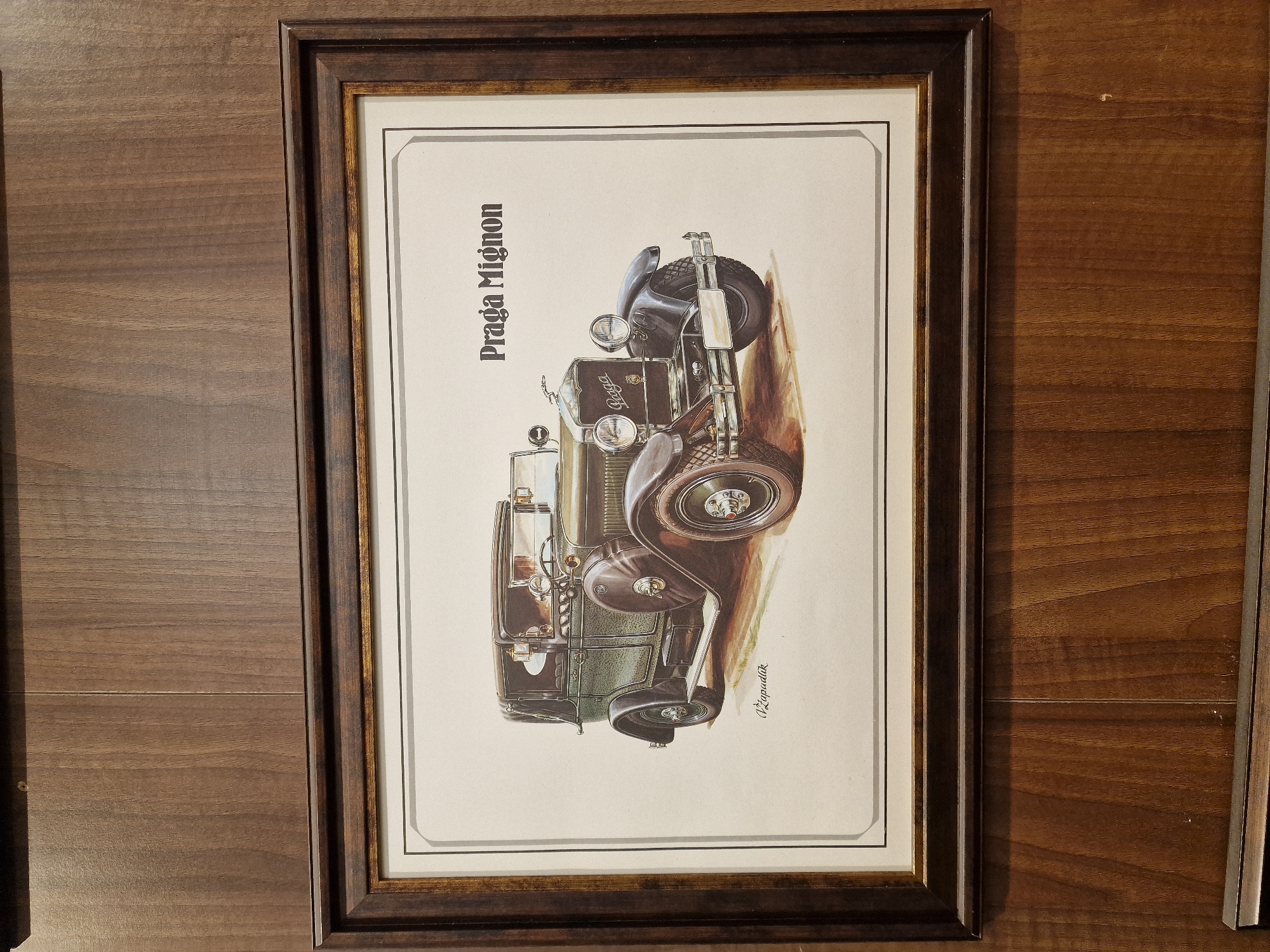 This is a framed print of a vintage car. The print features an illustration of a dark-colored car, possibly a roadster, with its wheels turned and the vehicle appearing to be in motion or posed on an incline. The car has a classic design with visible headlights, a grille, and spoked wheels. The artwork is rendered in a realistic style with a focus on shading and detail, giving it a somewhat aged appearance. The background of the print is a pale, off-white color, with a subtle watercolor effect at the bottom.
The print is framed in a dark wood frame with a simple, slightly beveled profile. The matting around the print is a cream color with a thin black border.
Text visible in the print includes "Praga Mignon" written vertically in a stylized font on the left side of the image, above the car. On the right side of the image, near the bottom, there is a signature that appears to read "V. Zapalák".
The overall impression is of a piece of automotive art, likely intended to commemorate or celebrate the "Praga Mignon" model. The image's orientation and the illustration style suggest it might be a reproduction of an older advertisement or a dedicated artistic rendering.