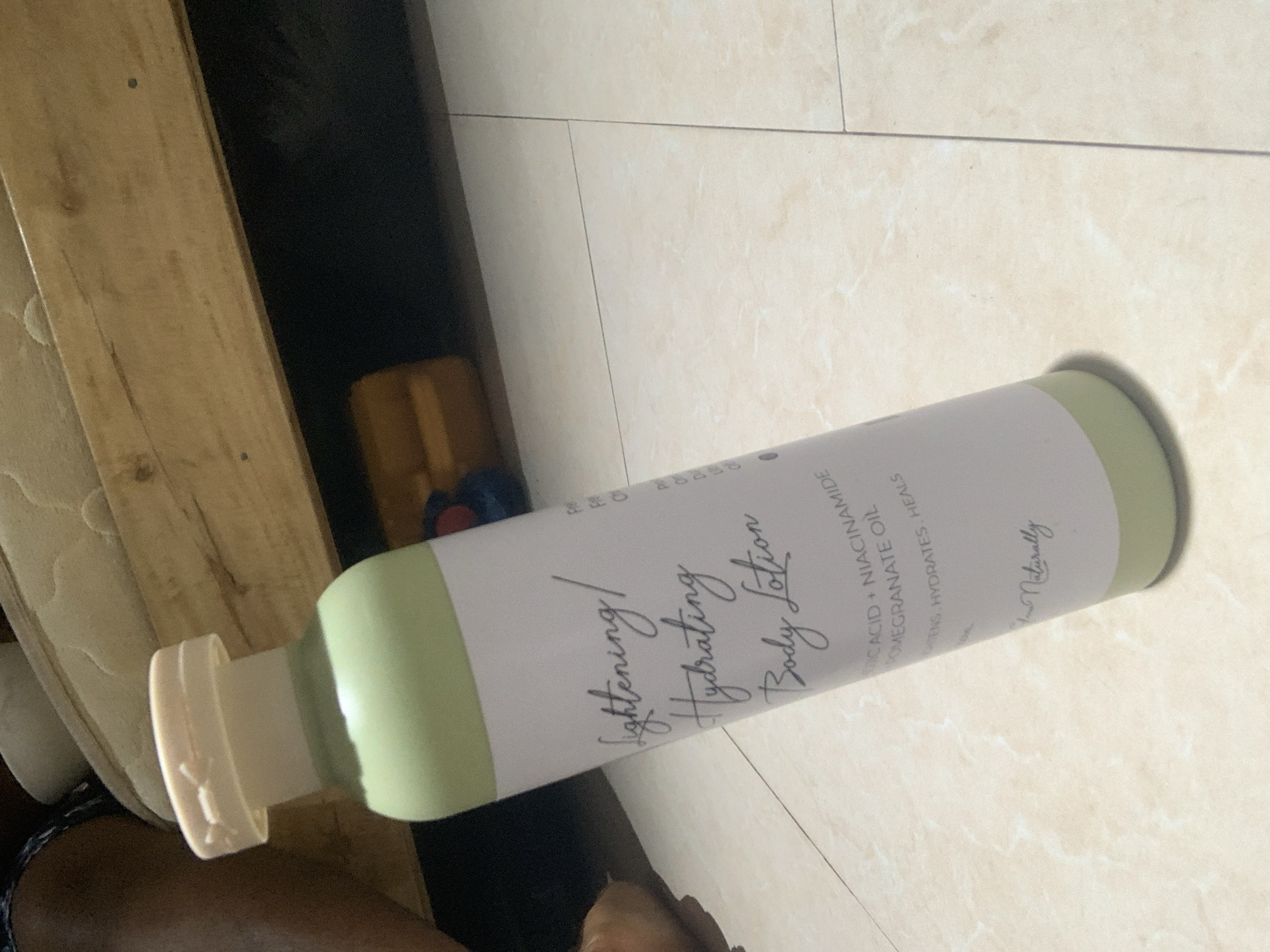 A light green and white bottle of "Lightening/Hydrating Body Lotion" with handwritten-style text is placed on a beige tiled floor, angled towards the viewer. The bottle has a light tan cap. Behind the bottle, a wooden structure, possibly a bed frame, is visible, with a mattress on top. The background is dark and out of focus, with a hint of a yellow object. The image is likely taken indoors, possibly in a residential setting. There is no indication of a specific location within Abuja, Nigeria.