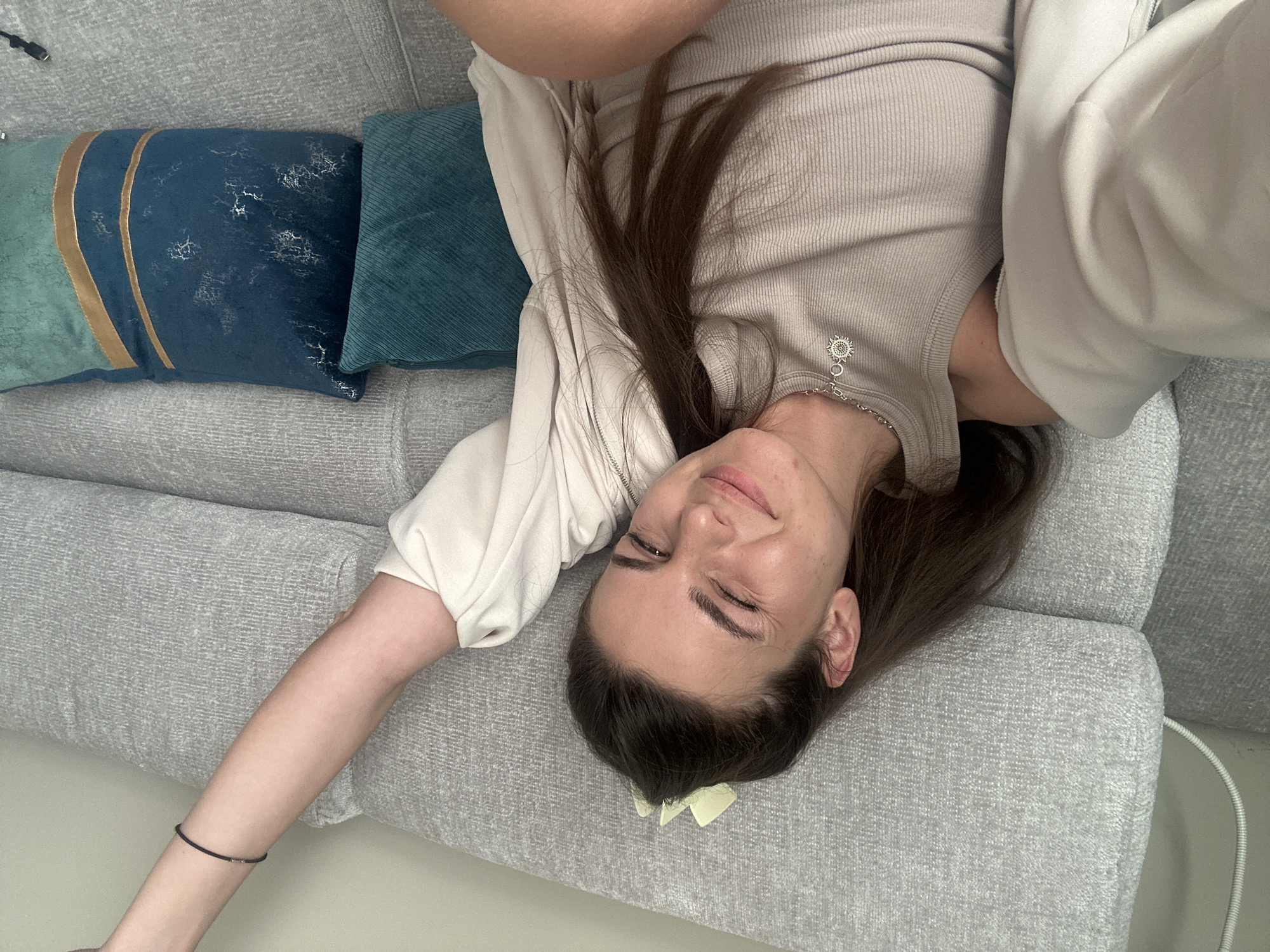 A woman is lying upside down on a gray couch. She has brown hair and is wearing a light-colored top and pants. There are two decorative pillows on the couch to her left. The location is likely a private residence; there is no indication that the image was taken in Plovdiv, Bulgaria.