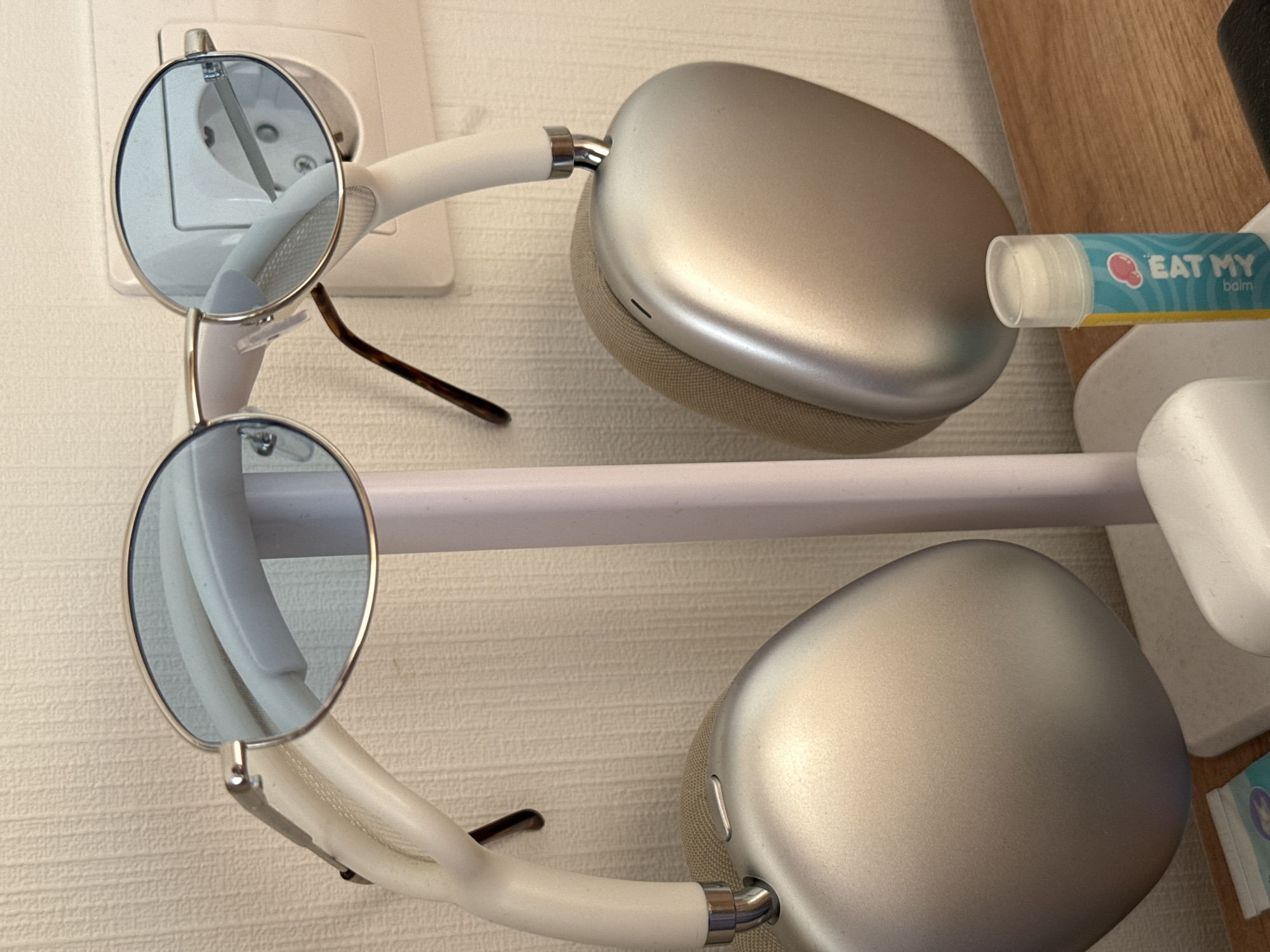 The image shows a close-up of a pair of silver Apple AirPods Max headphones, a pair of light blue tinted oval sunglasses, and a small tube of lip balm with the text "EAT MY balm" printed on it. The items are placed on a light-colored stand against a textured light-colored wall. A white electrical outlet is visible on the wall behind the headphones. The scene appears to be indoors, likely in a domestic setting. There is no indication of the city of Lyubertsy, Russia, in the image.