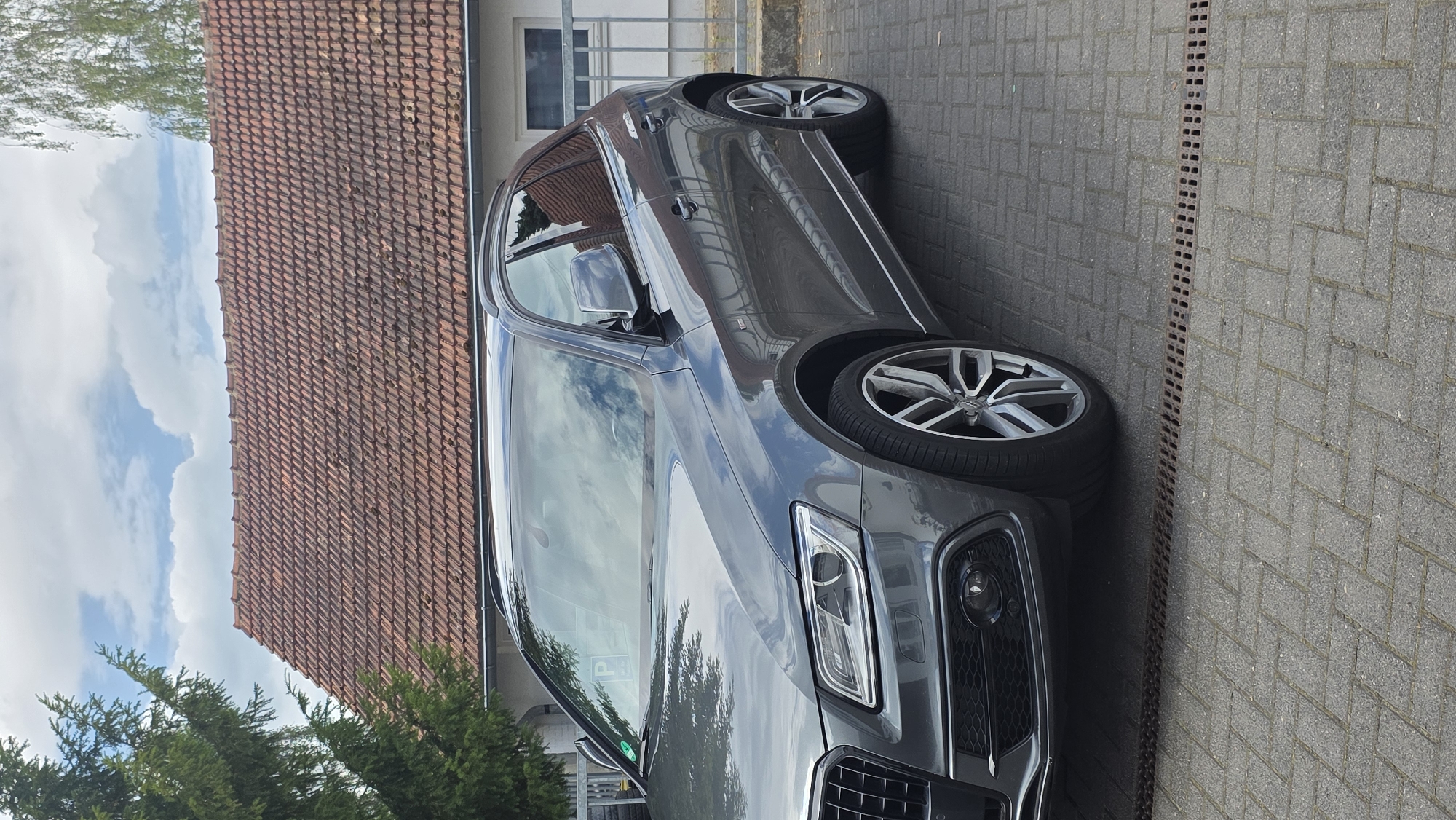 A dark gray Audi SUV is parked on a gray brick driveway. The car is angled towards the right side of the frame. The front of the car is on the right, and the rear is on the left. The car has silver rims.
To the left of the car is a red tile roof. Above the roof is a blue sky with white clouds. Green trees are visible in the lower left corner and along the left side of the roof. The background shows a building with a white exterior and a window. There is a drain running along the edge of the driveway.
The image is taken during the daytime, with bright natural light and shadows visible. The scene appears to be a residential or commercial area, possibly a driveway in front of a house or business. No people are visible in the image. There is a visible parking sign on the windshield of the car.