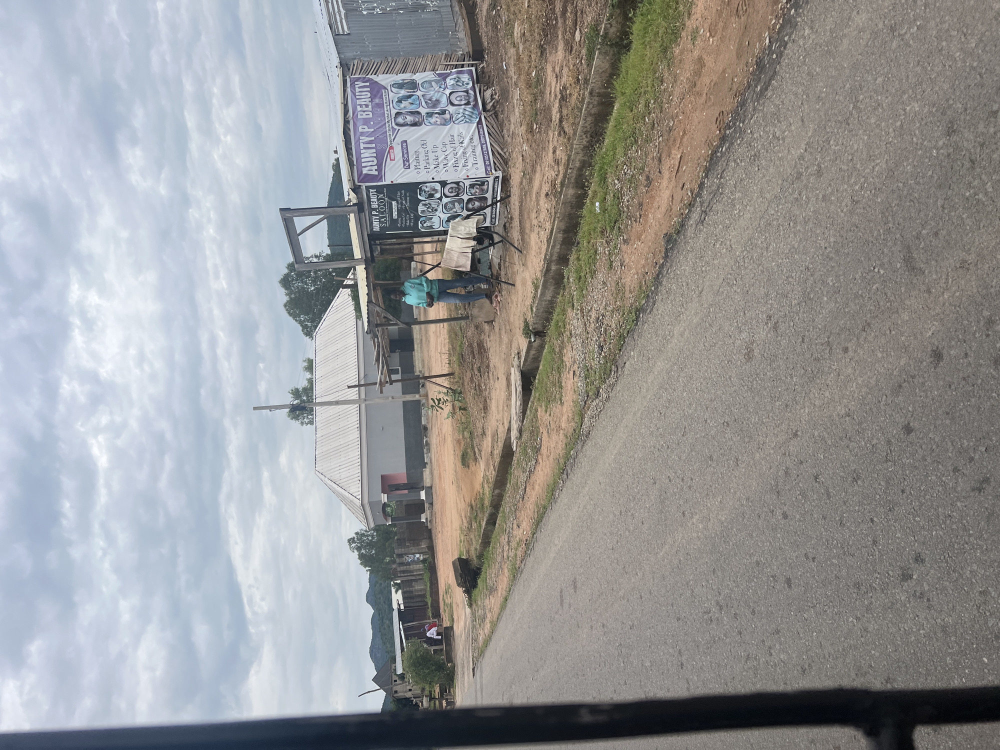 The image shows a street scene in Jalingo, Nigeria. A prominent sign for "Aunty P. Beauty Saloon" displays various hair and beauty services offered, including plaiting, makeup, and nail fixing, along with photos of hairstyles. The salon appears to be an open-air setup, with chairs and a person sitting outside, suggesting it's daytime. The surroundings include a dirt road, some greenery, and buildings in the background, including one with a corrugated metal roof. The sky is overcast with clouds. The overall impression is of a local business operating in a semi-urban or rural setting in Nigeria. The text on the sign provides contact information and a list of services, indicating a commercial activity.