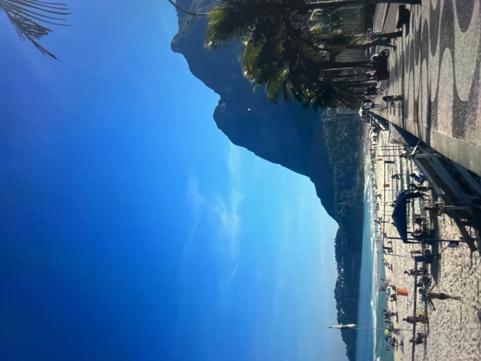 A wide shot captures a bustling beach scene in Rio de Janeiro, Brazil, with the iconic Sugarloaf Mountain in the background. The foreground features a curved boardwalk with a distinctive mosaic pattern, lined with palm trees. The beach is populated with numerous people sunbathing, playing sports, and relaxing under umbrellas. The ocean is visible with gentle waves, and a flag is planted on the sand. The sky is a clear, vibrant blue.