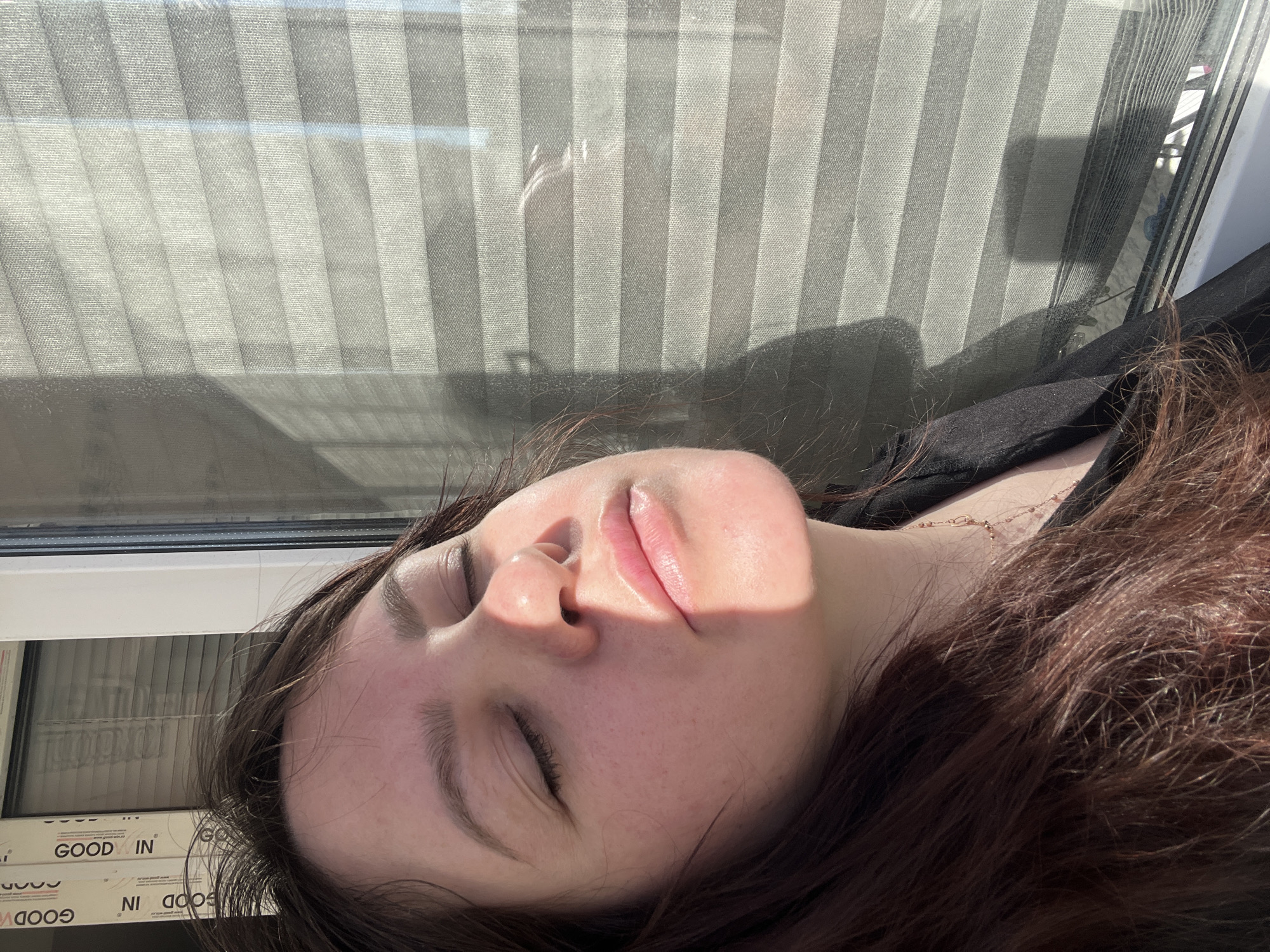 A fair-skinned woman with closed eyes and auburn hair rests near a window with closed blinds, likely in an indoor setting. Part of her face is brightly illuminated, while the rest of her head is in shadow. She is wearing a black garment and a delicate necklace is visible. The window frame has the words "GOOD IN" printed along the side. The city location in Noviy, Russia cannot be determined from the provided image.