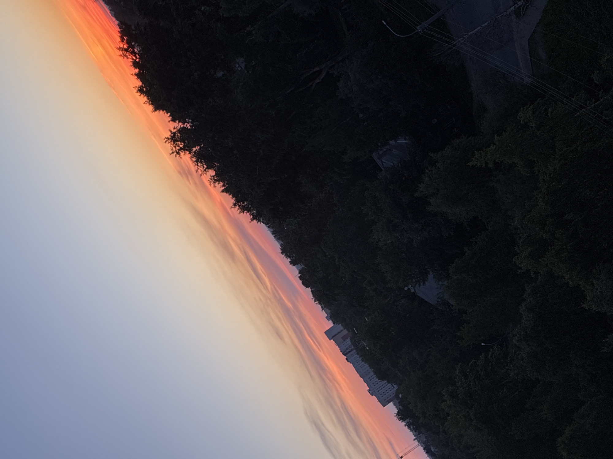 An aerial, diagonally tilted, medium shot captures a sunset over a suburban landscape. The sky transitions from a pale blue at the top left to vibrant orange and pink hues from a band of clouds stretching diagonally across the frame from the lower left towards the upper right. Below the sky, a dense canopy of dark green trees dominates the middle and right portions of the image, obscuring much of the ground. To the left, a row of tall, light-colored apartment buildings is visible peeking through the trees. A construction crane is also visible in the far left of the image, partially obscured by the trees. The lower right of the image shows a dark, paved area with visible power lines stretching across it. The overall scene suggests a residential area at dusk.