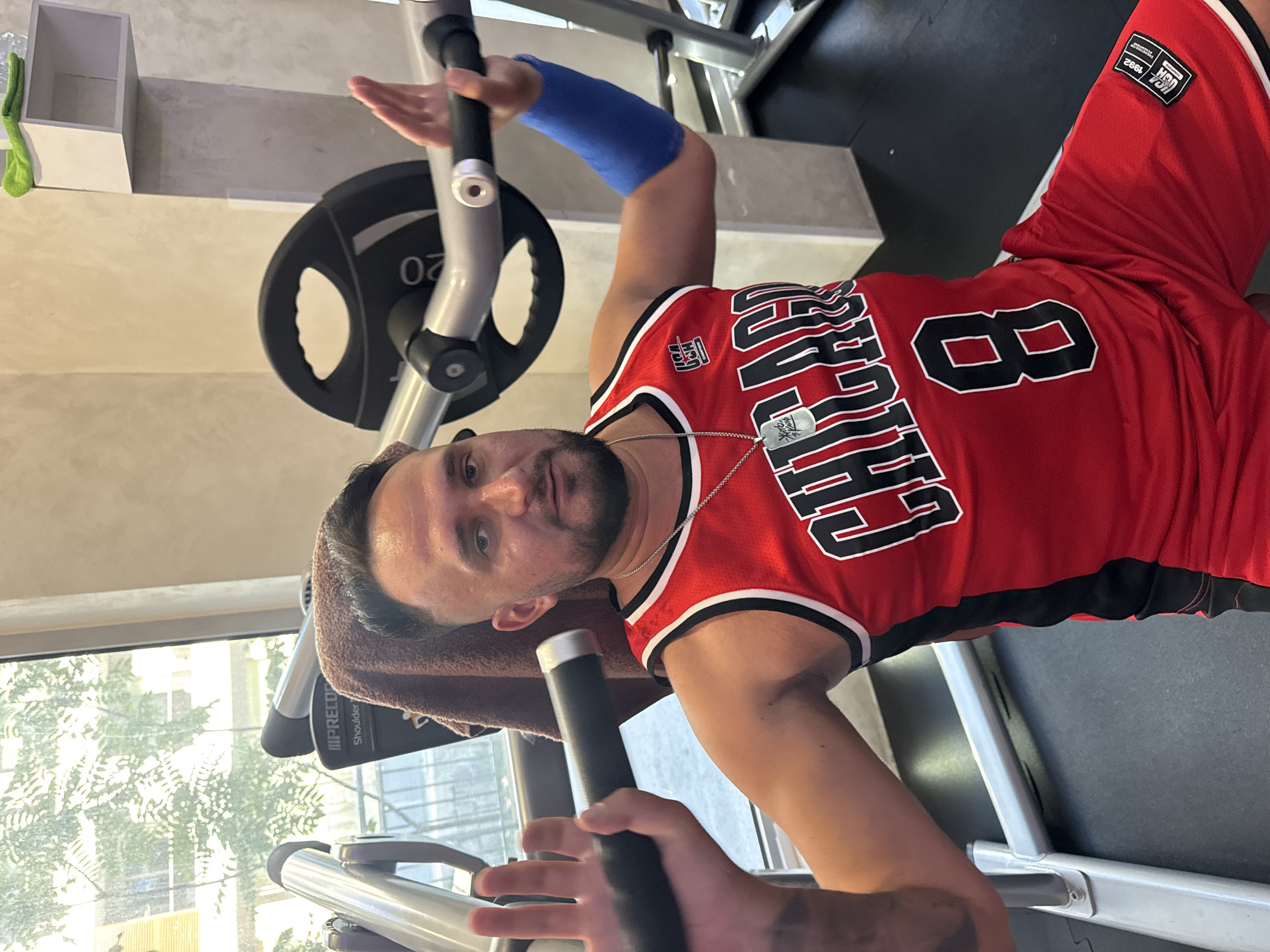 A man is pictured in a gym, wearing a red Chicago Bulls basketball jersey and shorts with the number 8. He has a blue compression sleeve on his right arm. The man is sitting on a weight machine, holding onto the bar with his hands. A brown towel is draped over the back of the machine behind his head. The gym appears to be indoors, with a window showing trees and a building outside. The overall setting suggests a fitness facility in Budva, Montenegro, given the context provided.