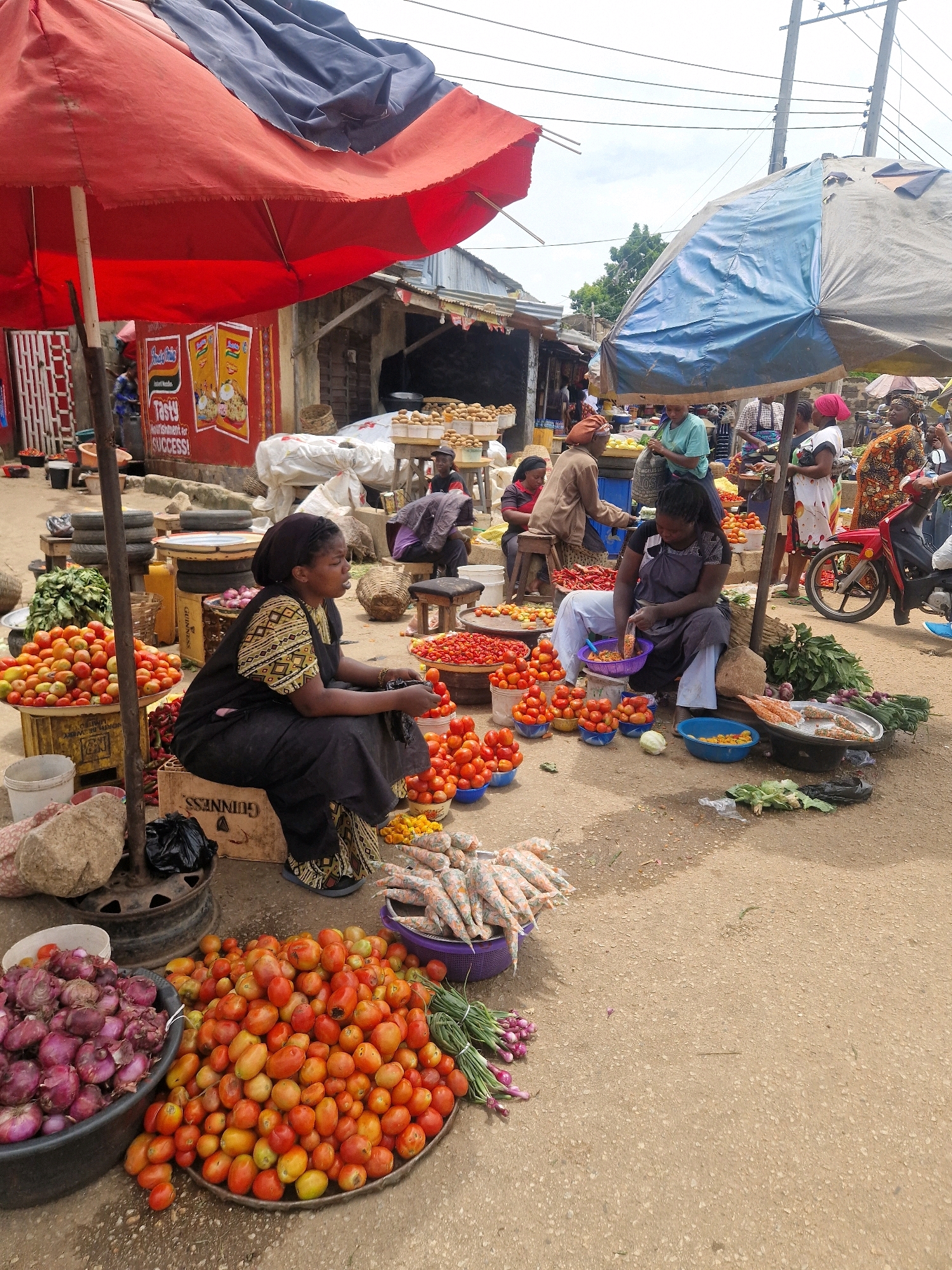 The image depicts an outdoor market scene in Kabong, Nigeria. A woman, wearing a black headscarf and a patterned dress, sits on a wooden crate next to a large display of tomatoes. She is looking to her right and appears to be talking. In front of her are bowls of tomatoes and what appear to be peppers. To her left, a large pile of red onions is in a black plastic basin. Behind the woman, various market stalls and other people are visible, along with a red motorcycle parked on the right side of the frame. The ground is paved with gravel and sand. The overall atmosphere suggests a typical bustling Nigerian market.